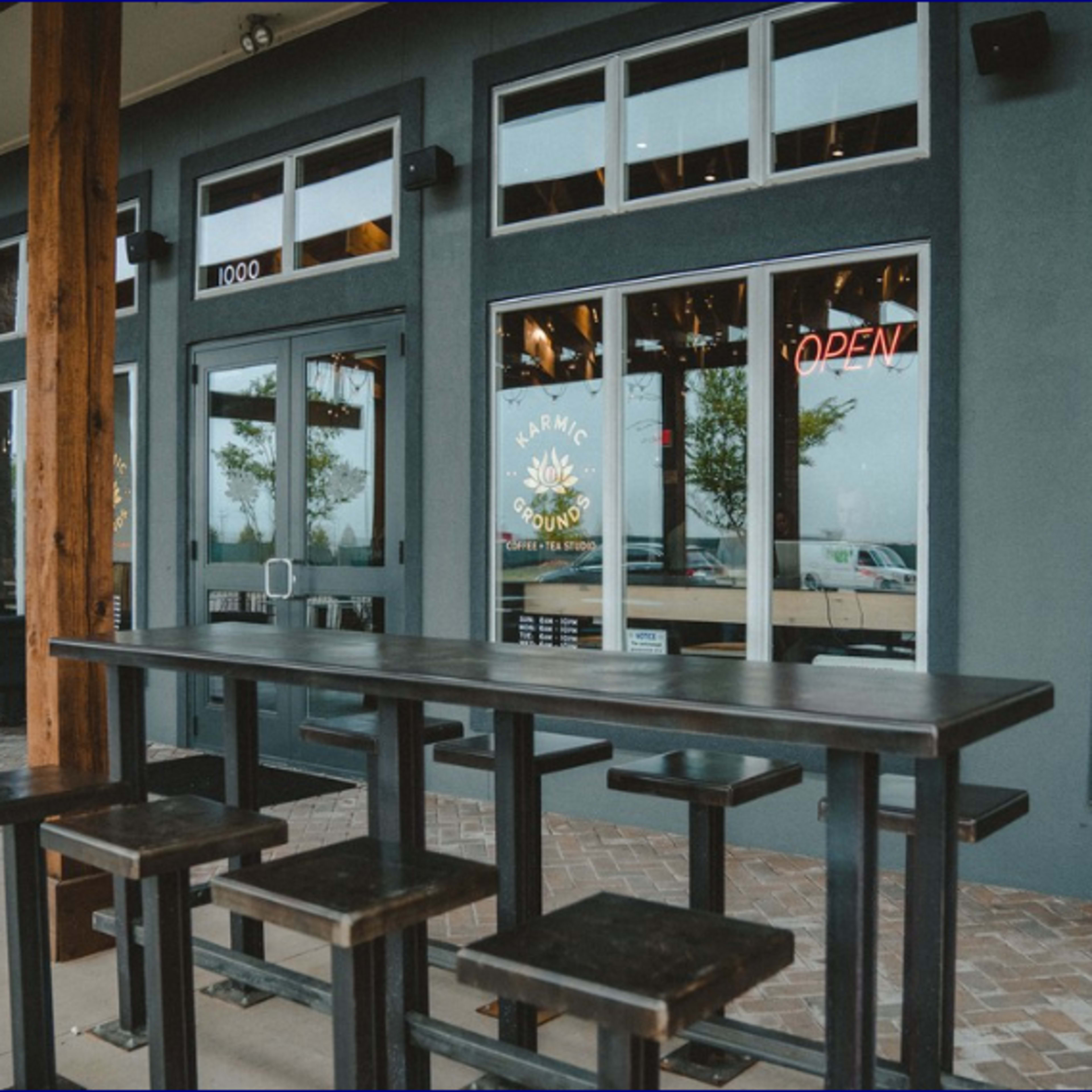 An outdoor seating area with a tall table and stools in front of a restaurant, featuring large windows and an "OPEN" sign.