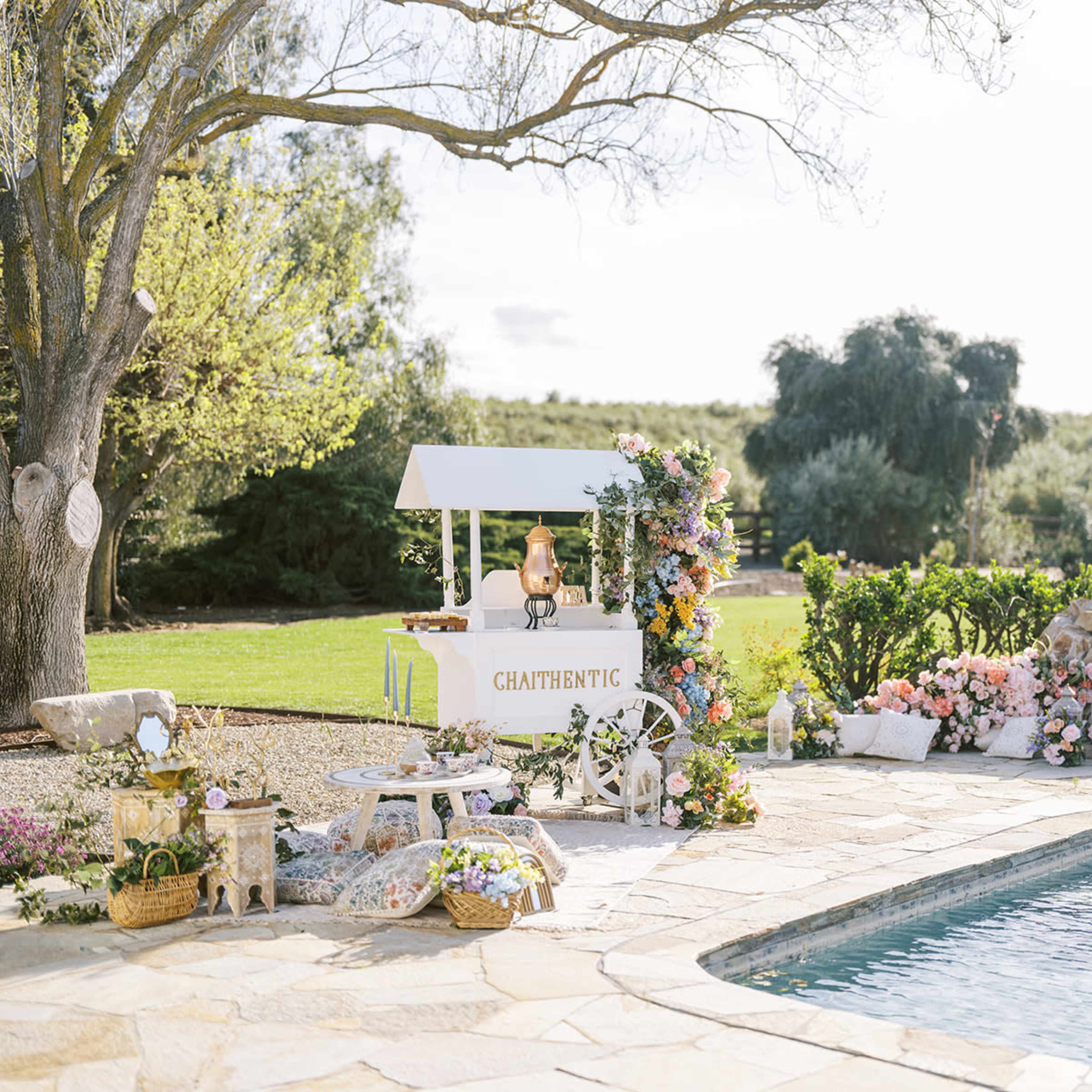 A white cart decorated with flowers stands beside a pool in a landscaped garden, with seating and various floral arrangements surrounding it.