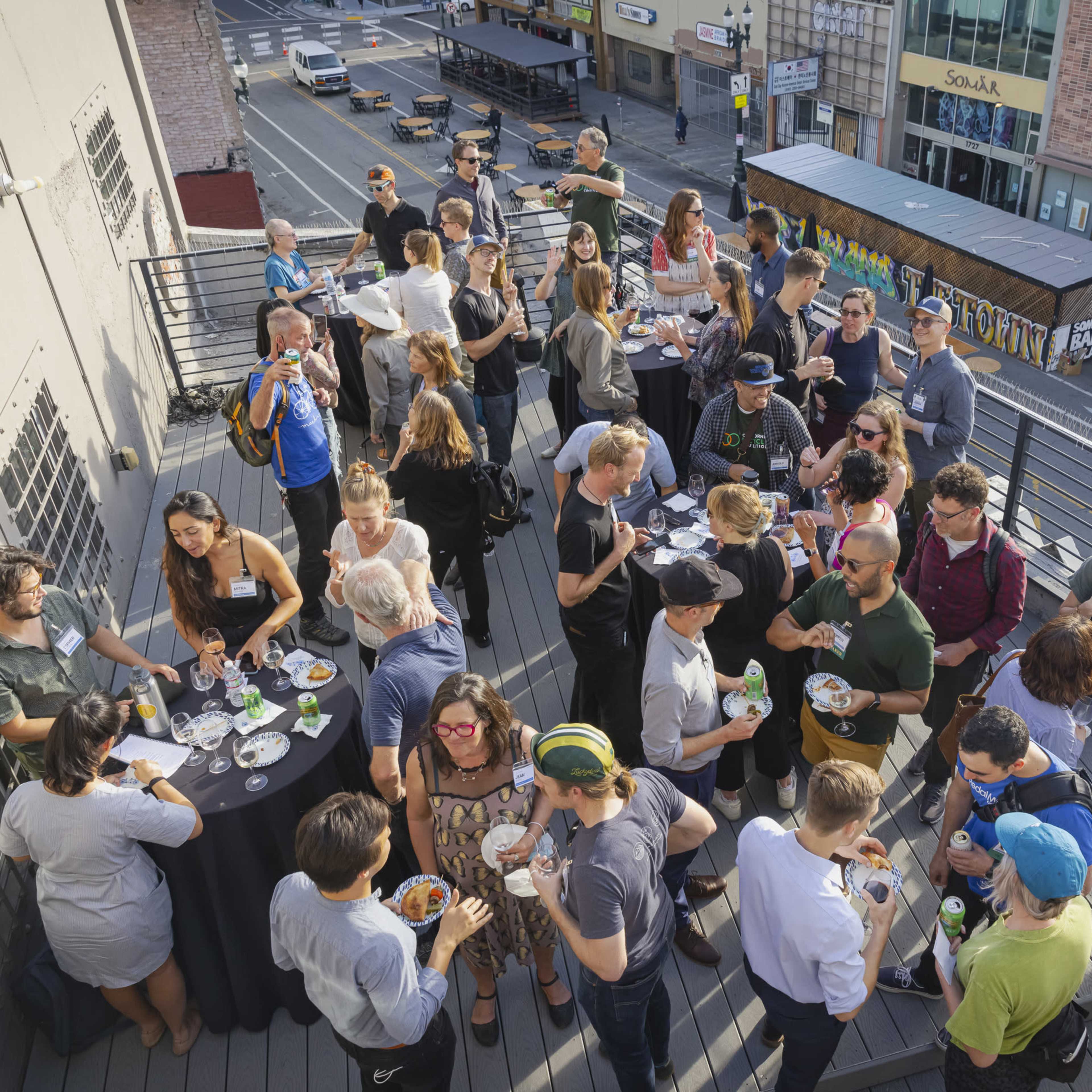 A large group of people socializes on an outdoor deck, enjoying food and drinks at an event in an urban setting.
