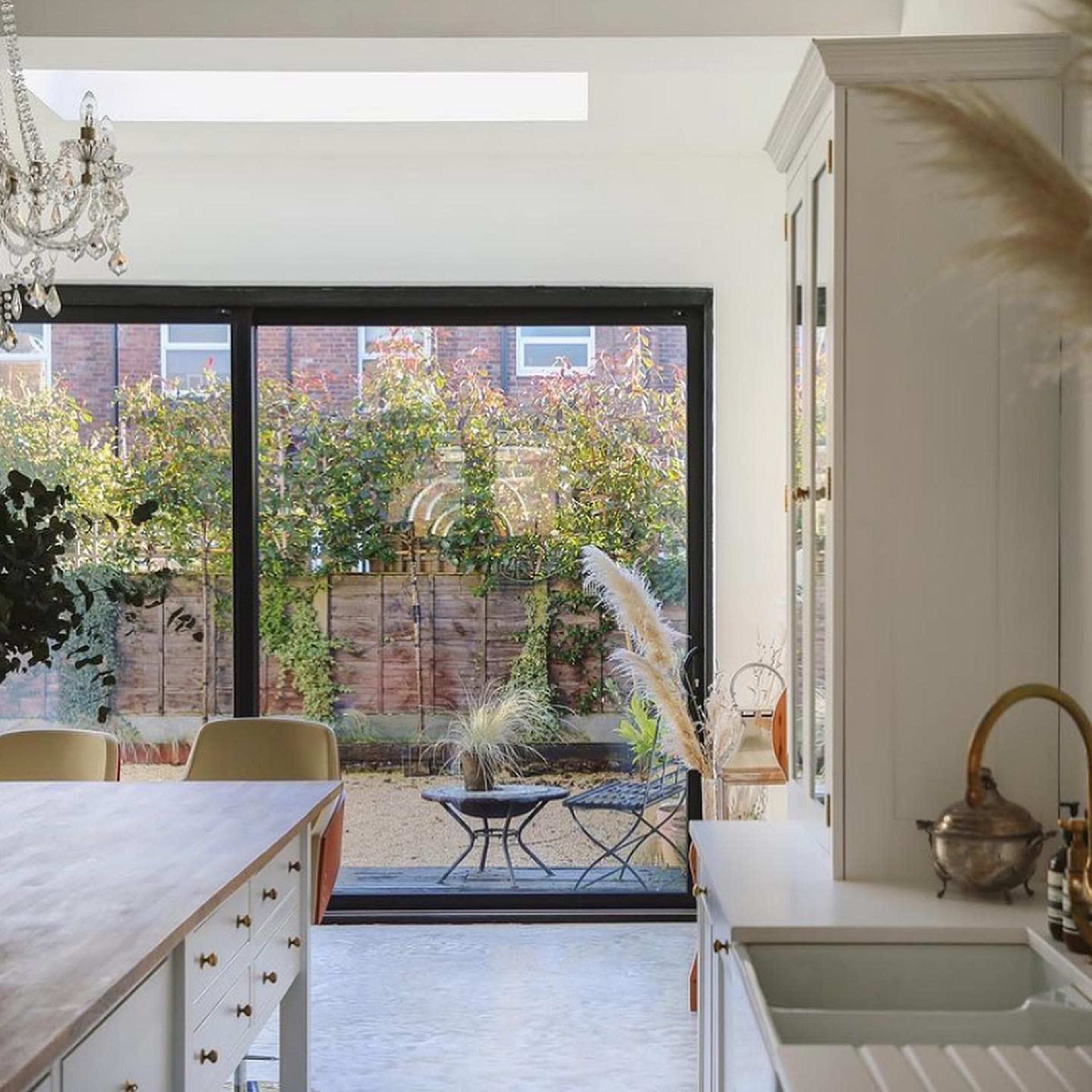 A modern kitchen with a large wooden island and a glass wall overlooking a garden with greenery and a patio set.