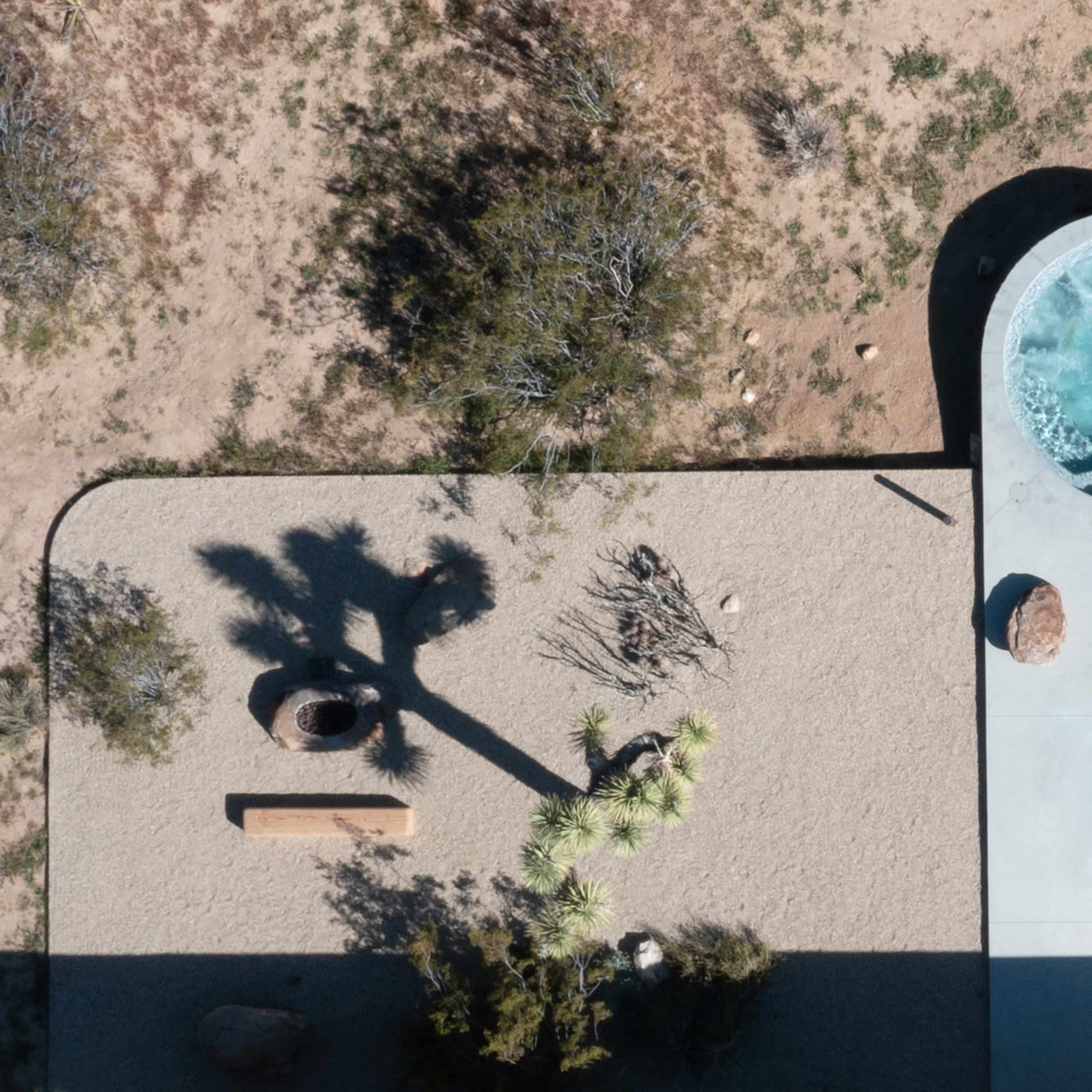 An aerial view shows a landscaped area with a circular hot tub, a stone bench, and sparse vegetation, including a palm tree casting a shadow, set against a sandy ground.