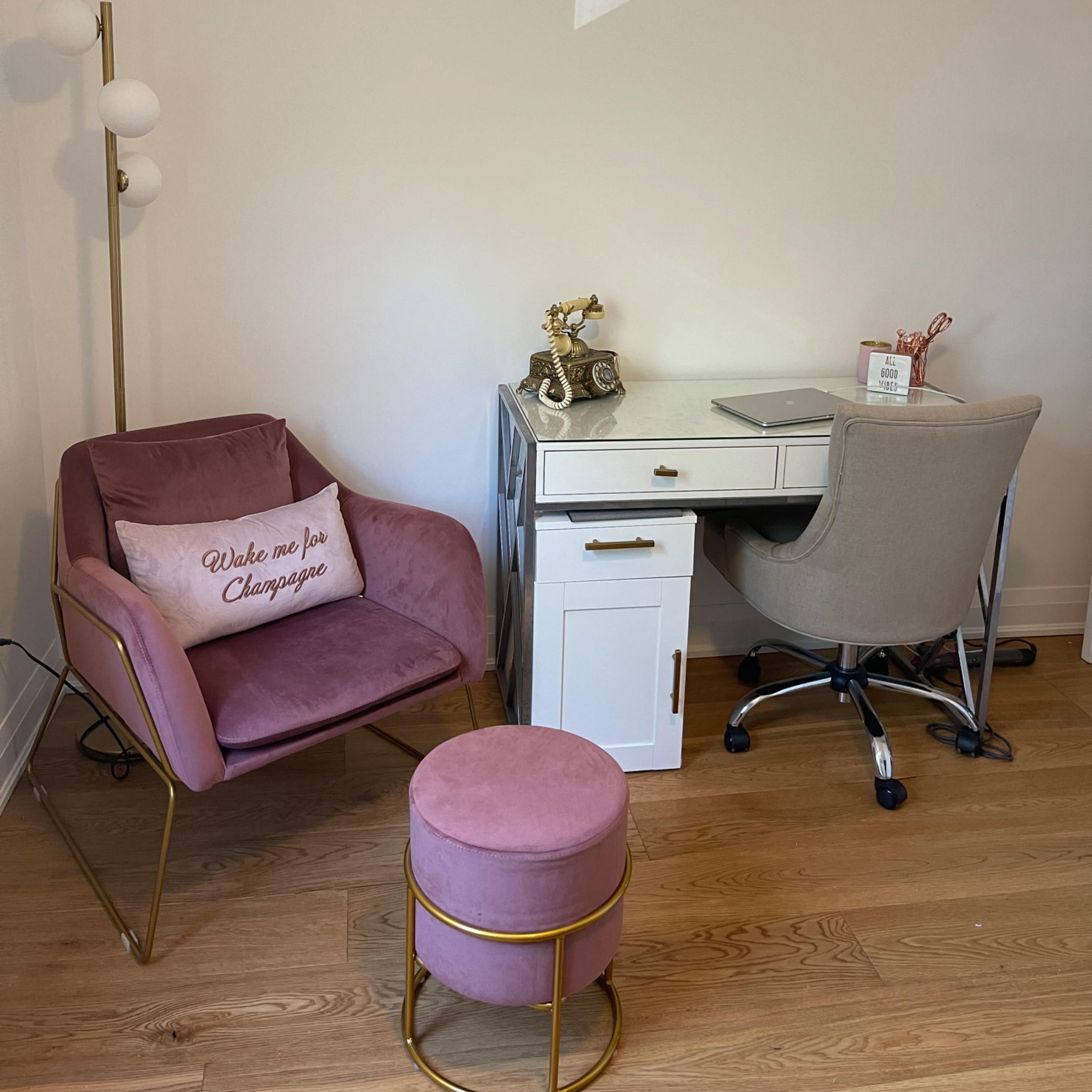 A cozy workspace featuring a white desk with a chair, a pink upholstered armchair, a small pink stool, and a floor lamp in a well-lit room.