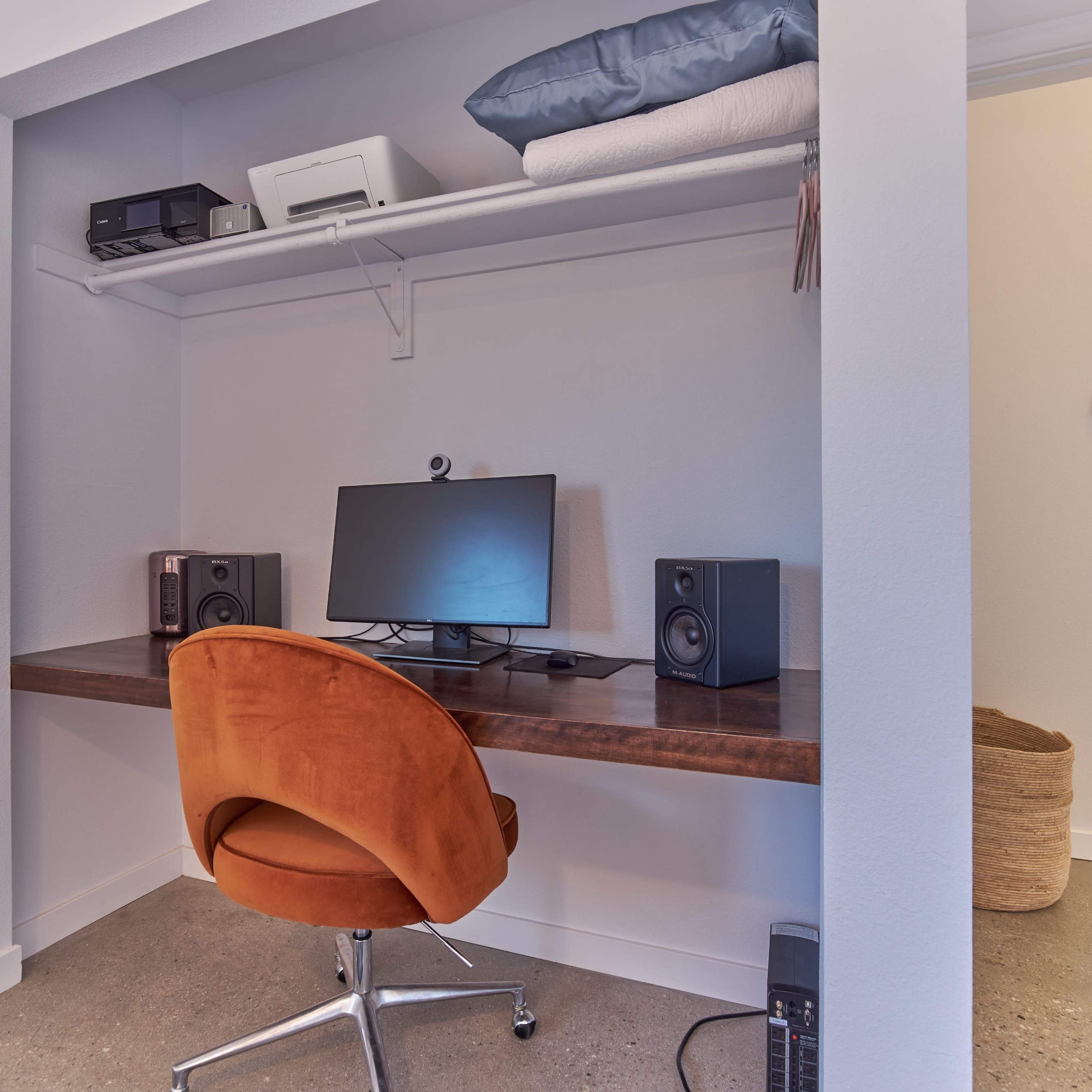 A small home office setup with a wooden desk, a computer monitor, speakers, and a comfortable orange swivel chair against a white wall.