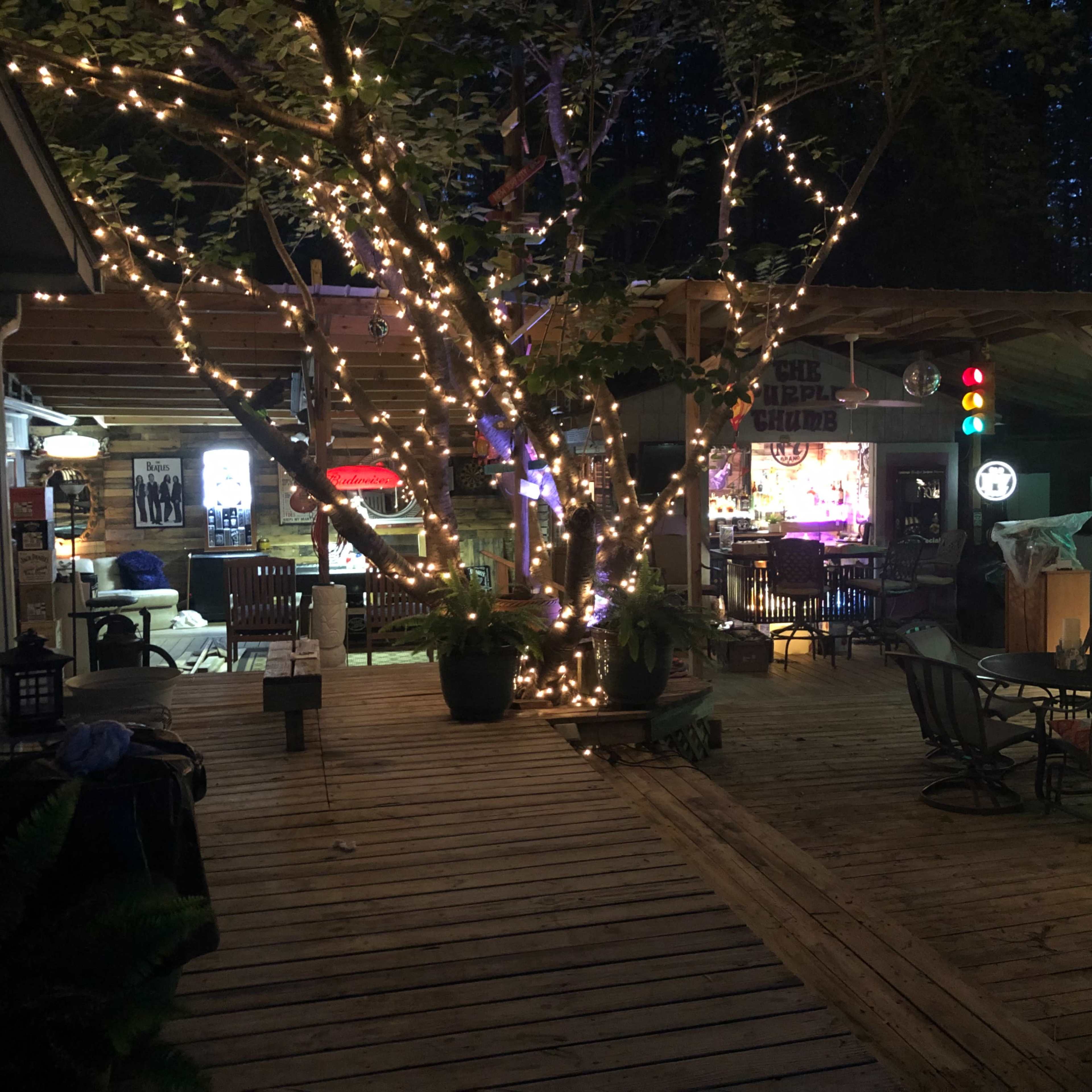 A wooden deck is adorned with string lights and features seating areas alongside a tree, creating a cozy outdoor space.