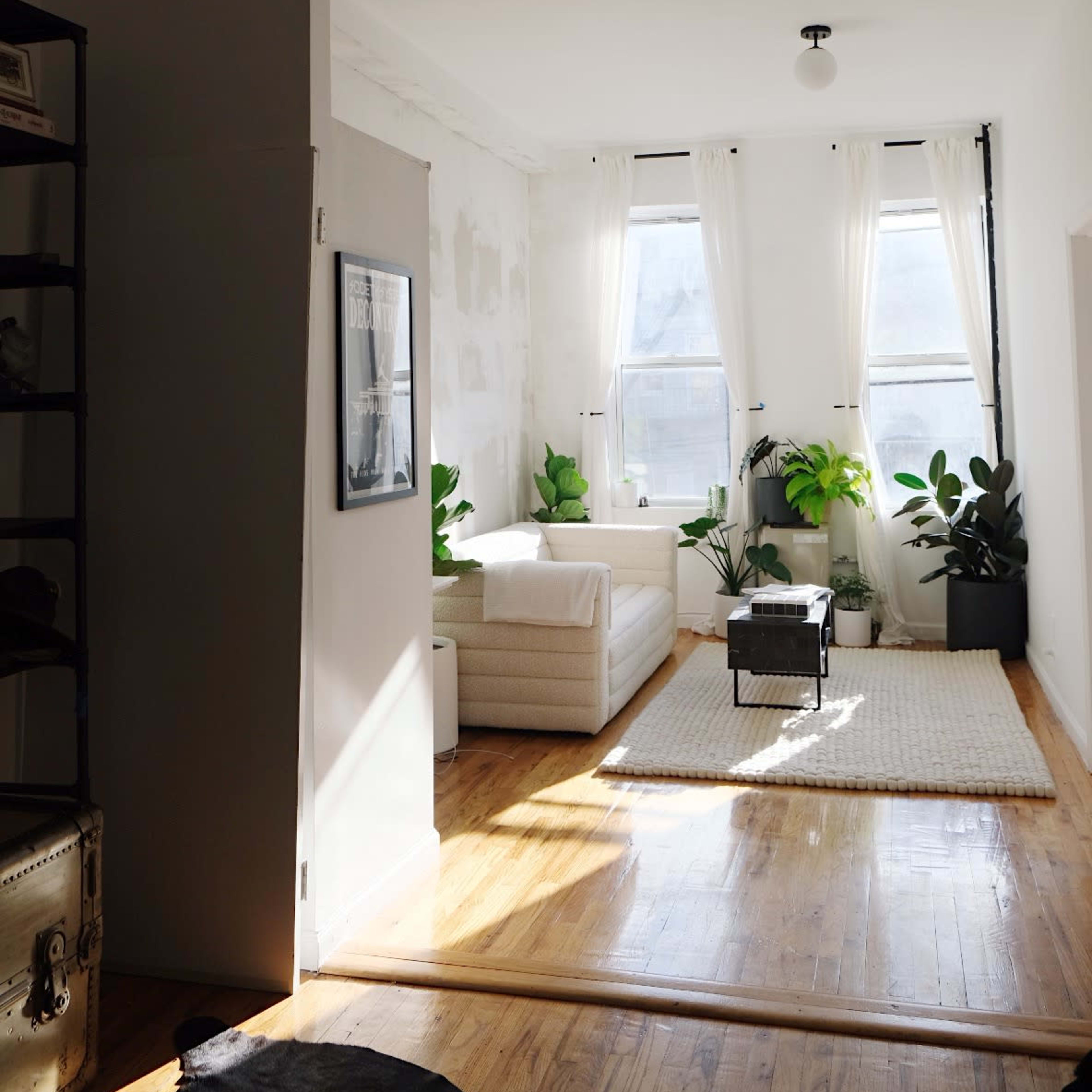 A bright, minimalistic room features large windows with sheer curtains, a white sofa, indoor plants, and a woven area rug on a wooden floor.