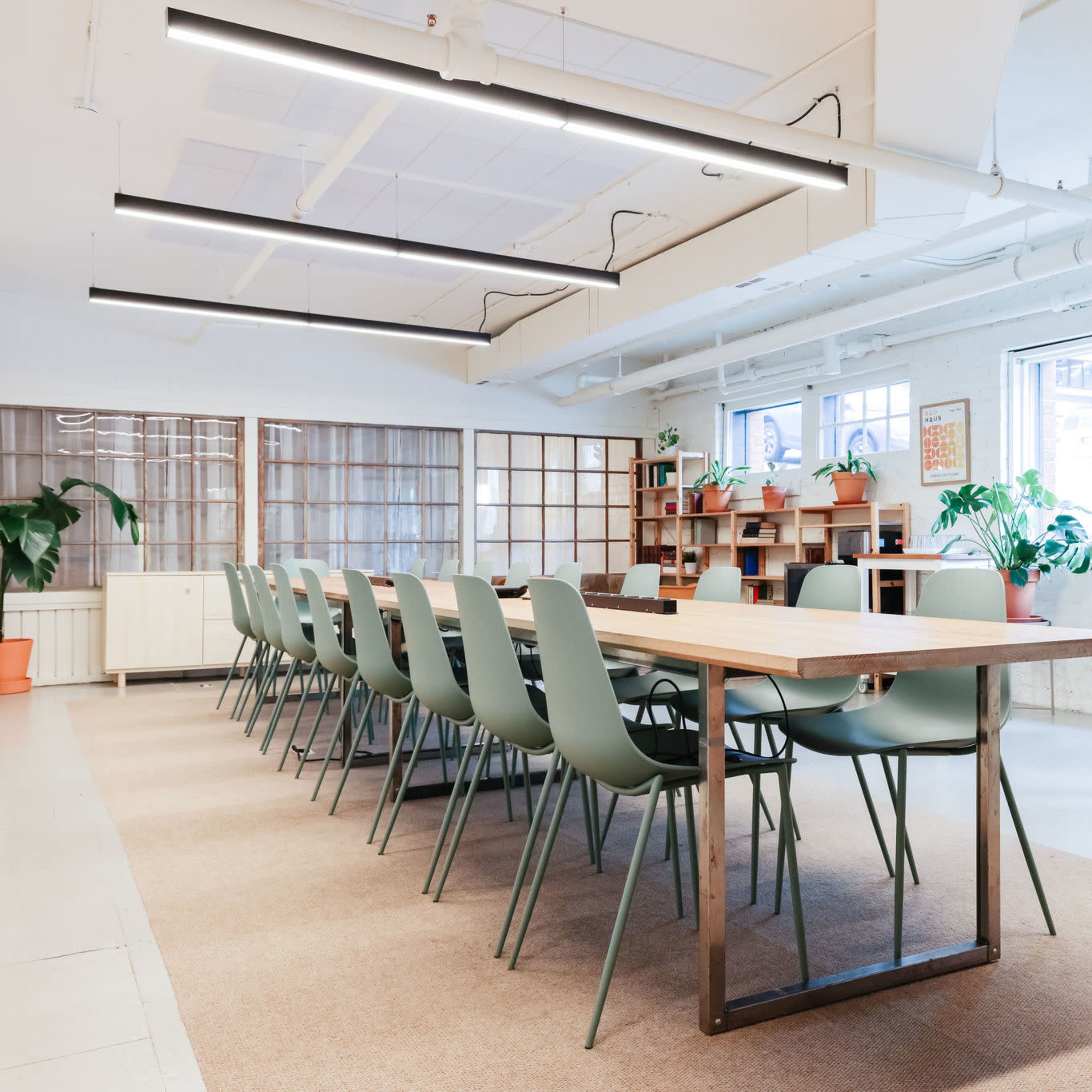 A modern conference room features a long wooden table surrounded by green chairs, with large windows and decorative plants in the background.