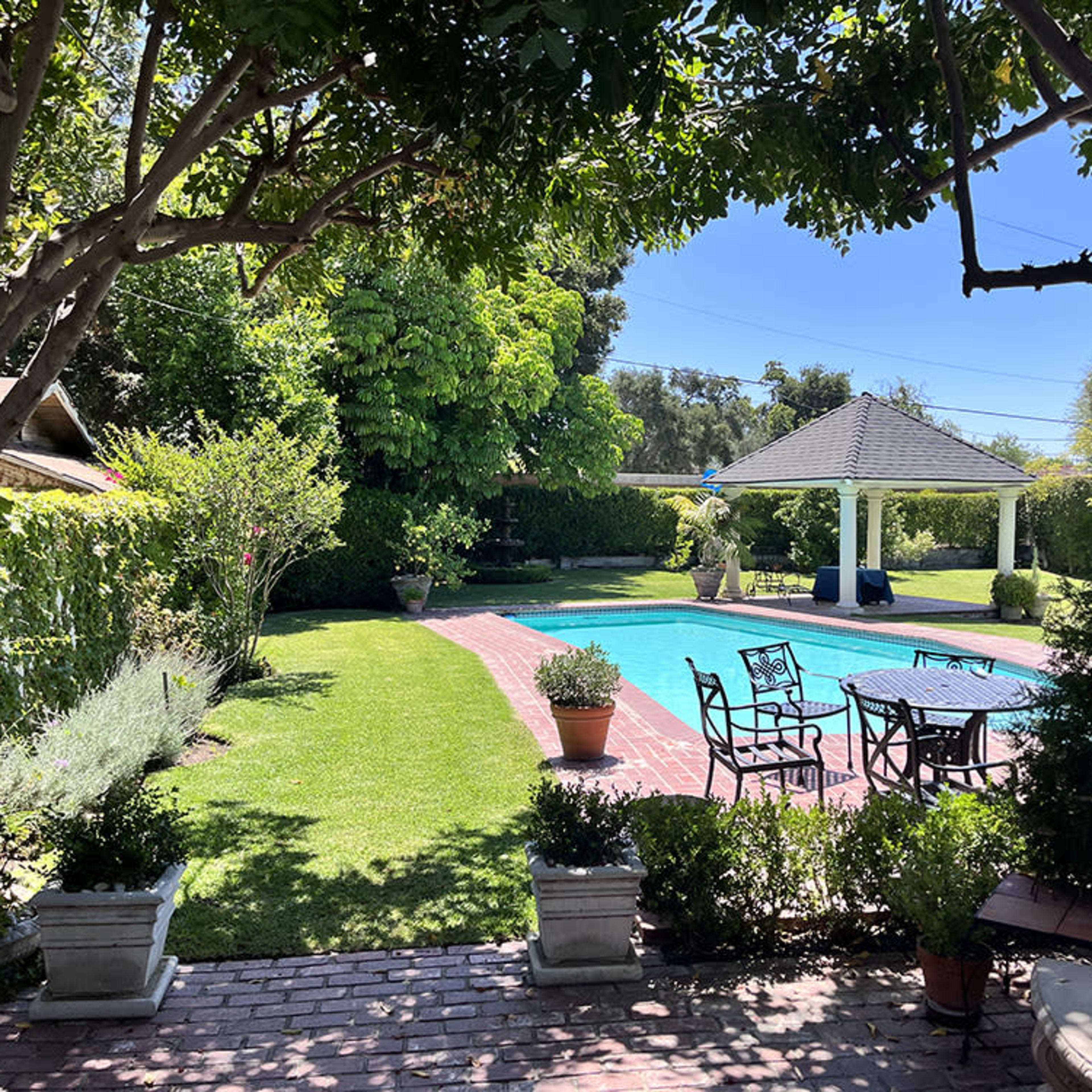 A well-maintained swimming pool is surrounded by landscaped greenery and patio furniture in a sunny backyard.