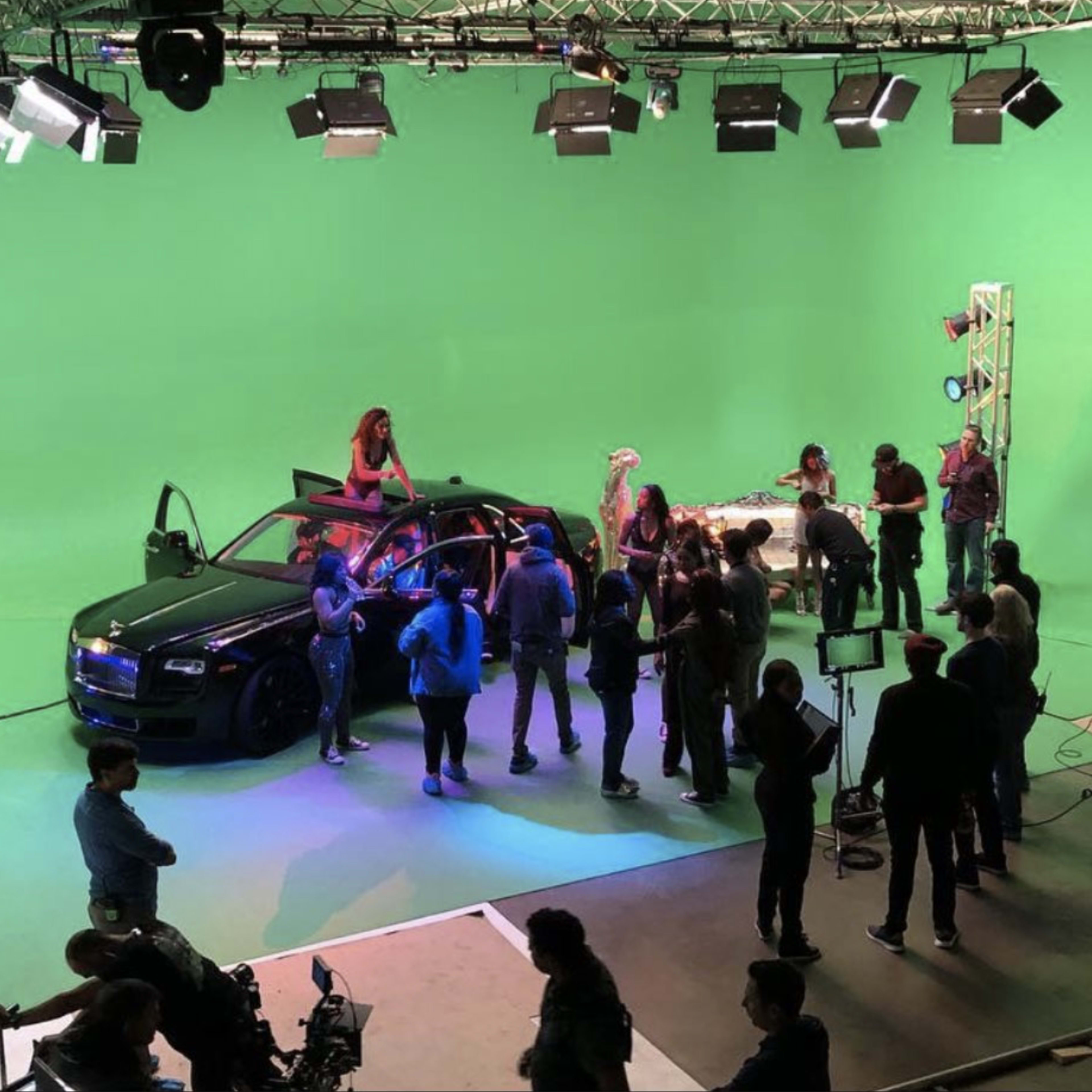 A group of people is gathered around a black car on a green screen set, with crew members and equipment visible in the background.