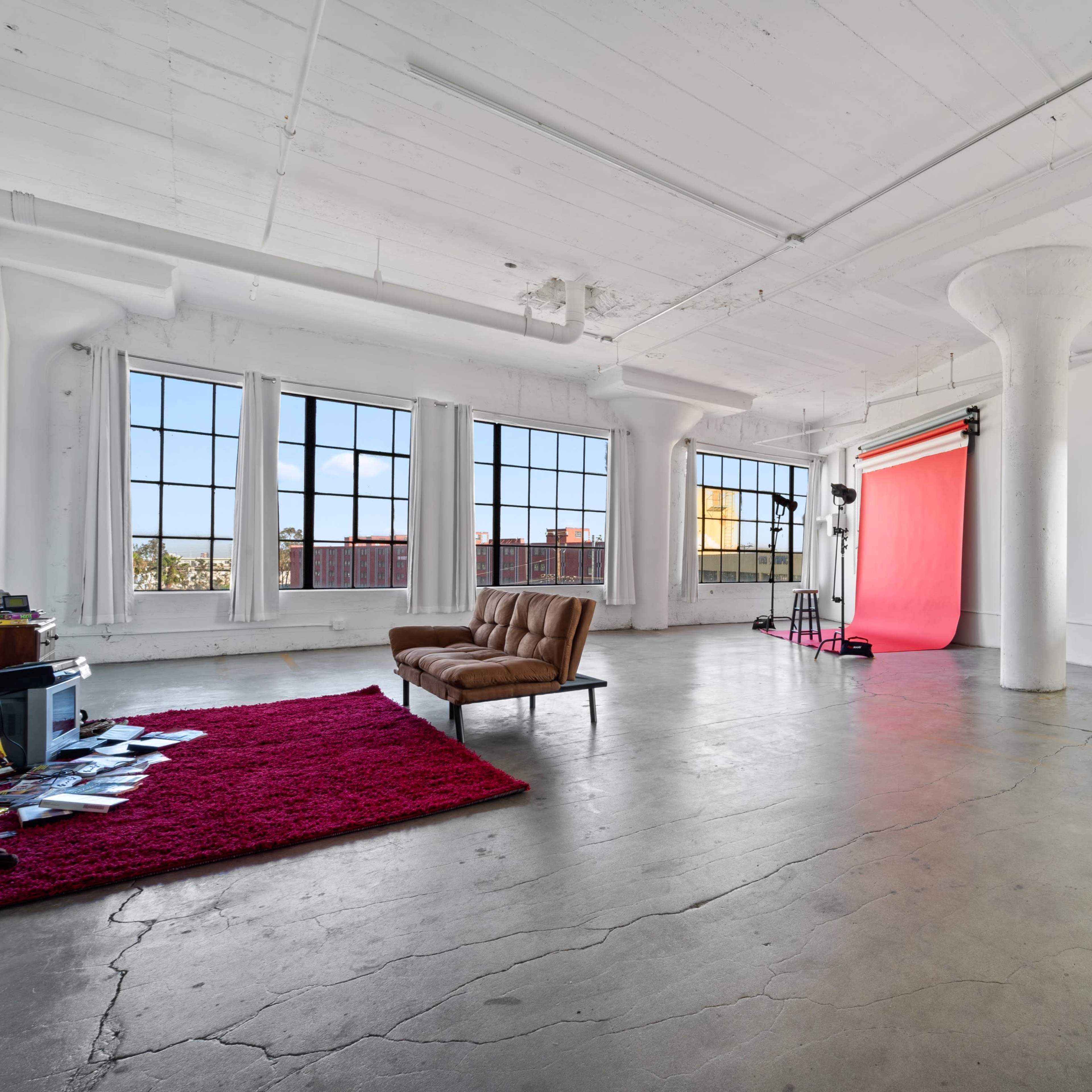 A spacious room with large windows, a brown sofa, a red rug, and a pink backdrop set up for photography.
