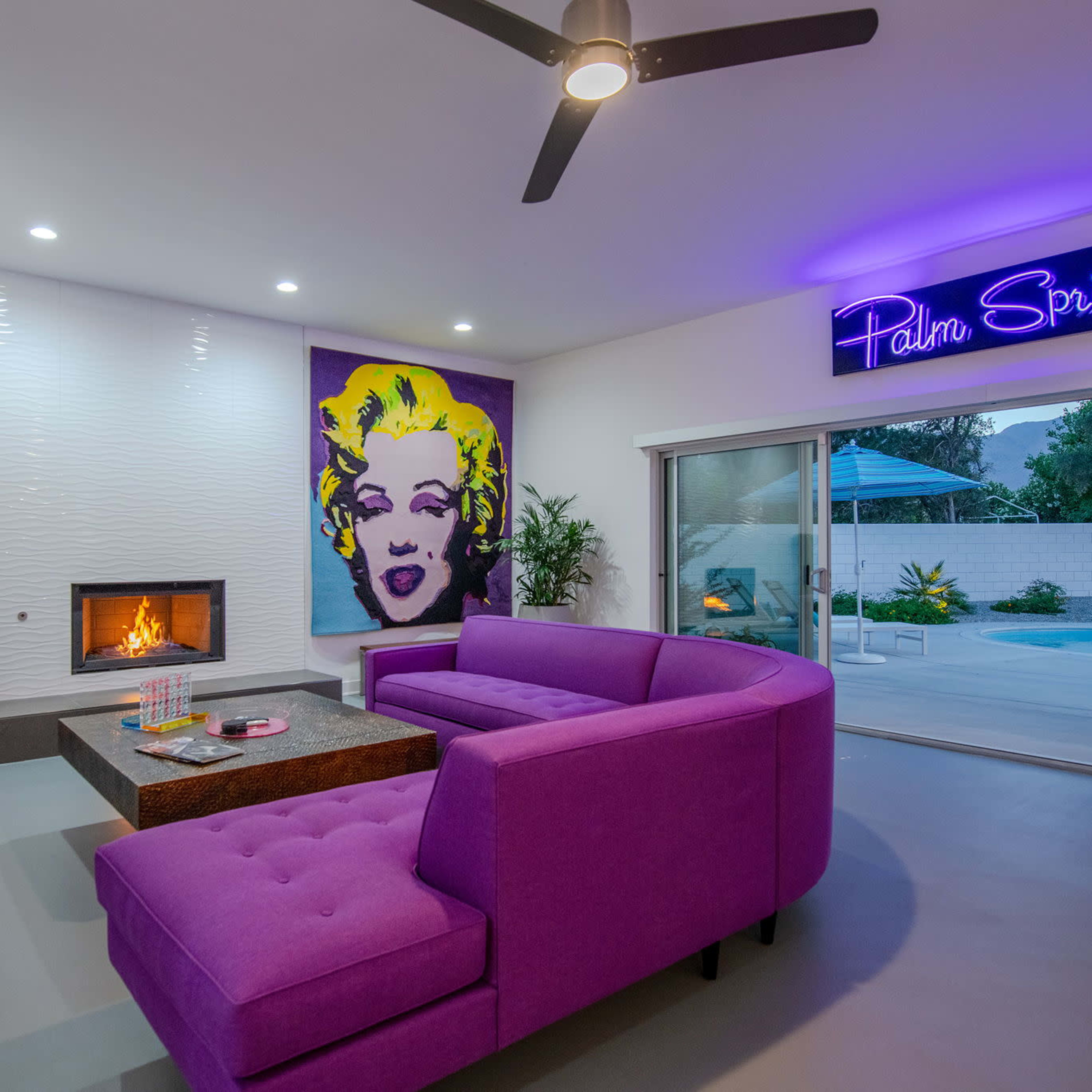 This interior space features a purple sectional sofa, a modern fireplace, and a large portrait of Marilyn Monroe against a light wall, with a neon "Palm Springs" sign and a black bear sculpture nearby.