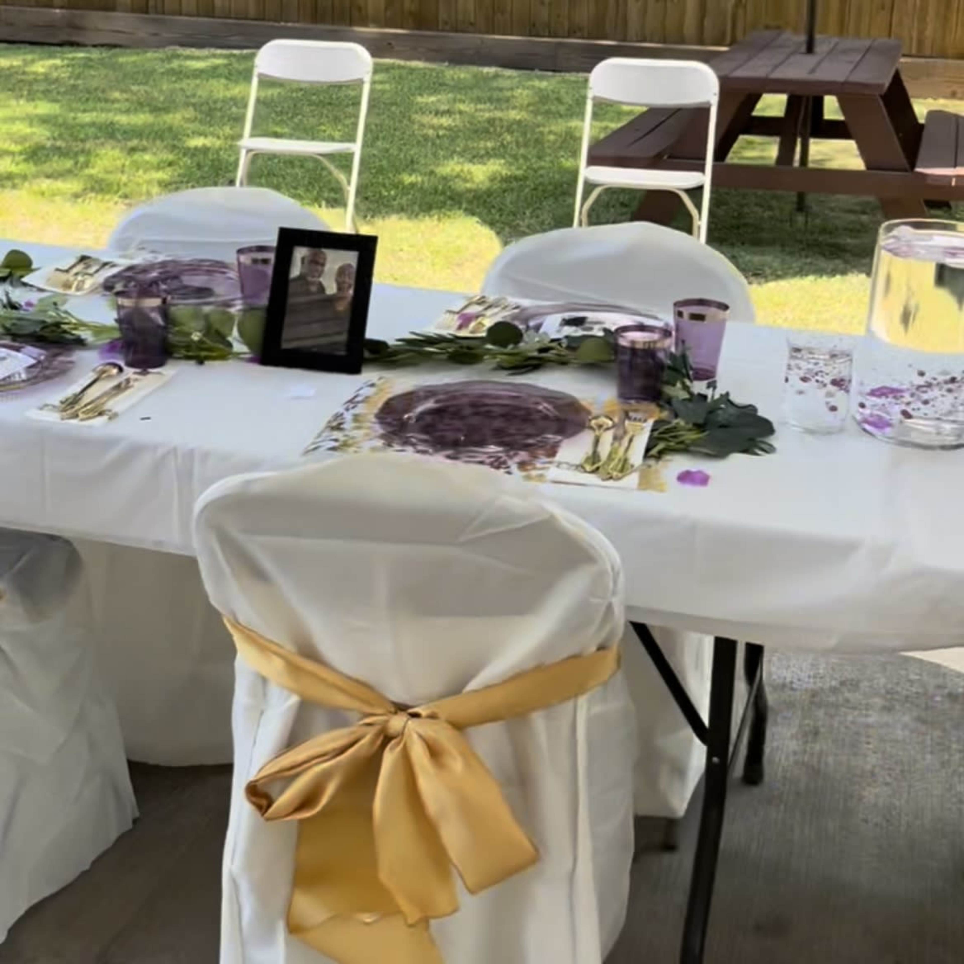 A table set for an outdoor gathering features white tablecloths, gold accents, clear glasses, and framed photographs surrounded by green grass and a shaded seating area.