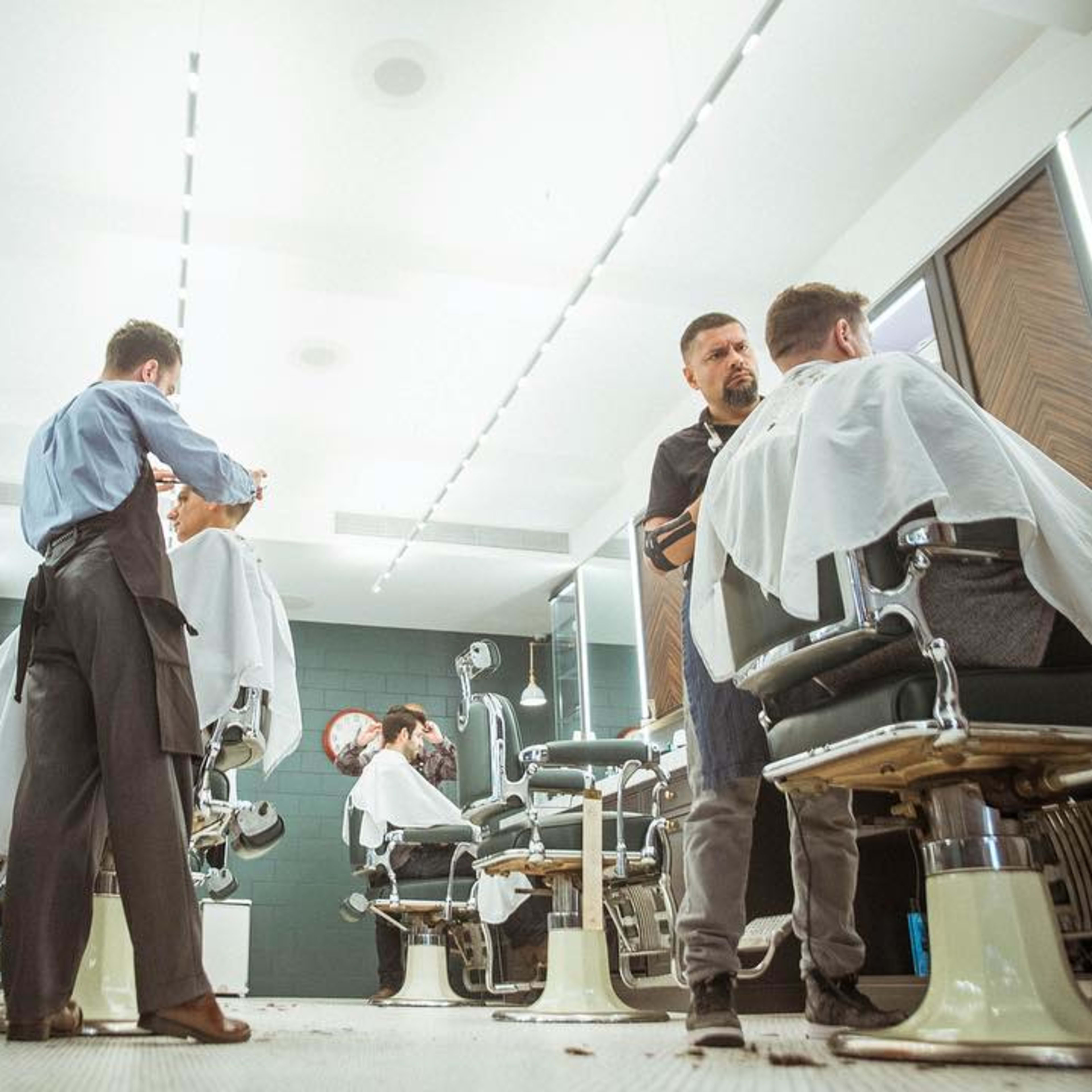 Two barbers attend to clients in a modern barbershop with stylish barber chairs and bright lighting.