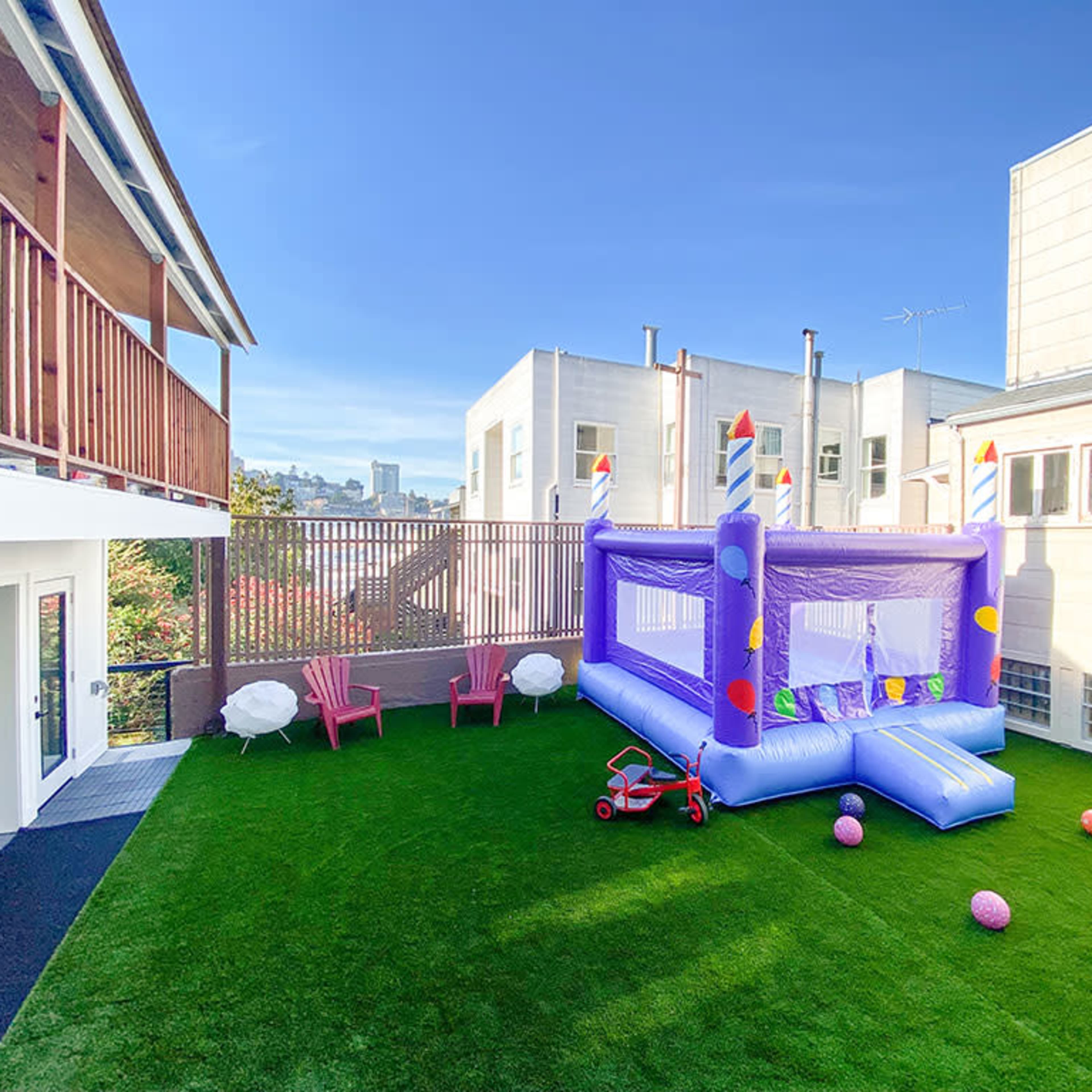 A colorful inflatable bounce house set up in a neatly maintained backyard with artificial grass, surrounded by lounging chairs and playground balls.