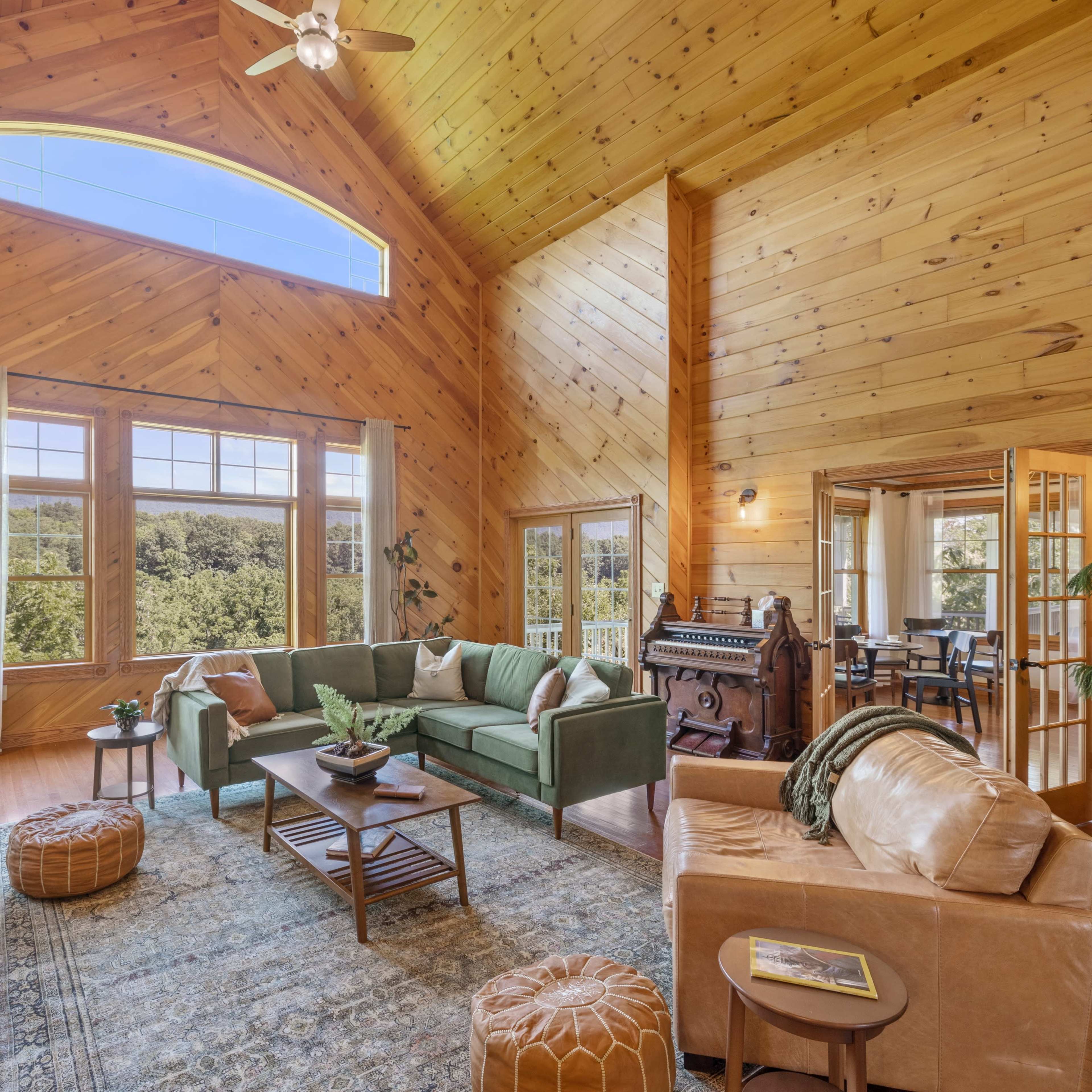 The room features a high ceiling with wooden beams, large windows showcasing a view of the outdoors, and a mix of comfortable seating arrangements.
