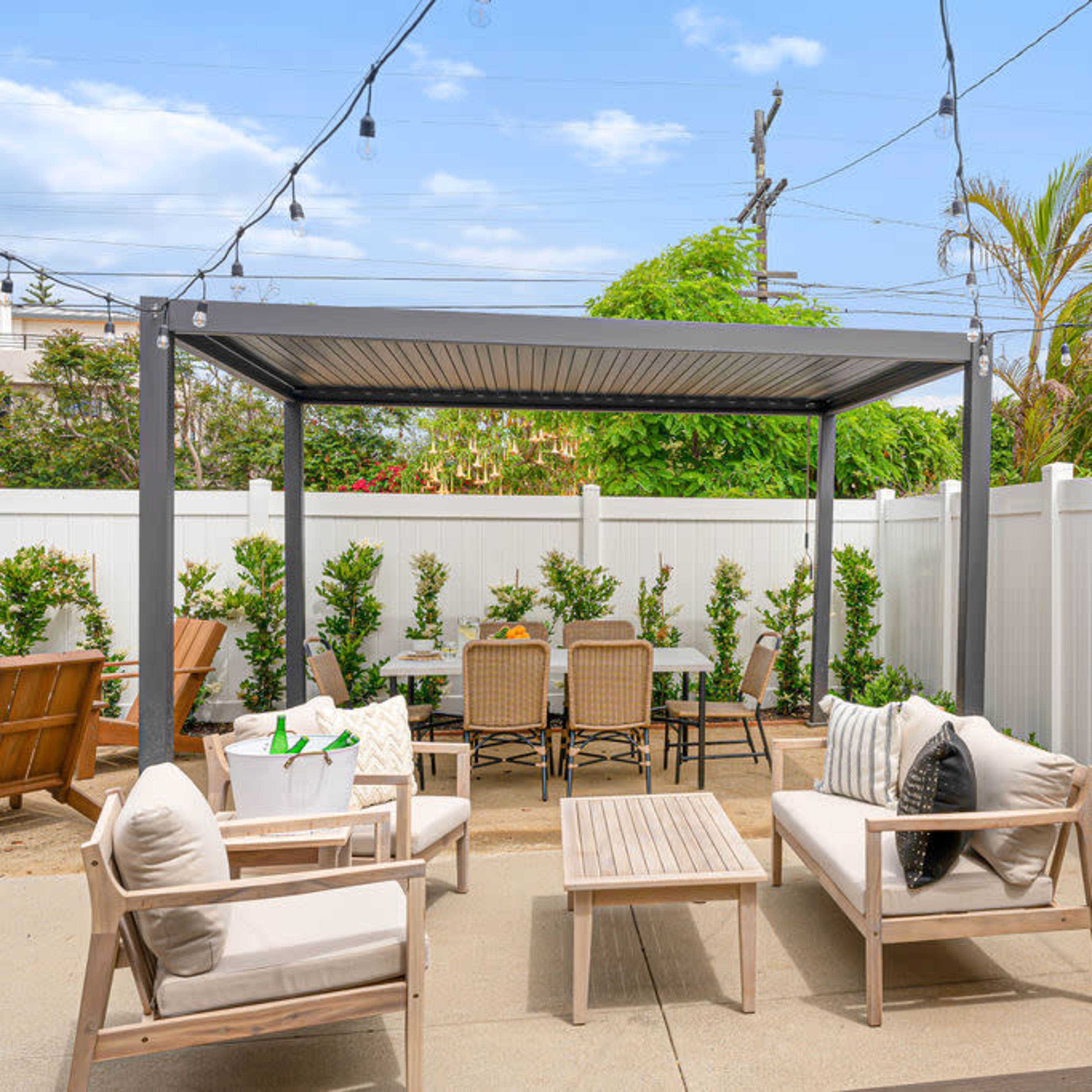 A patio area with a pergola, outdoor seating, a dining table, and greenery against a white fence.