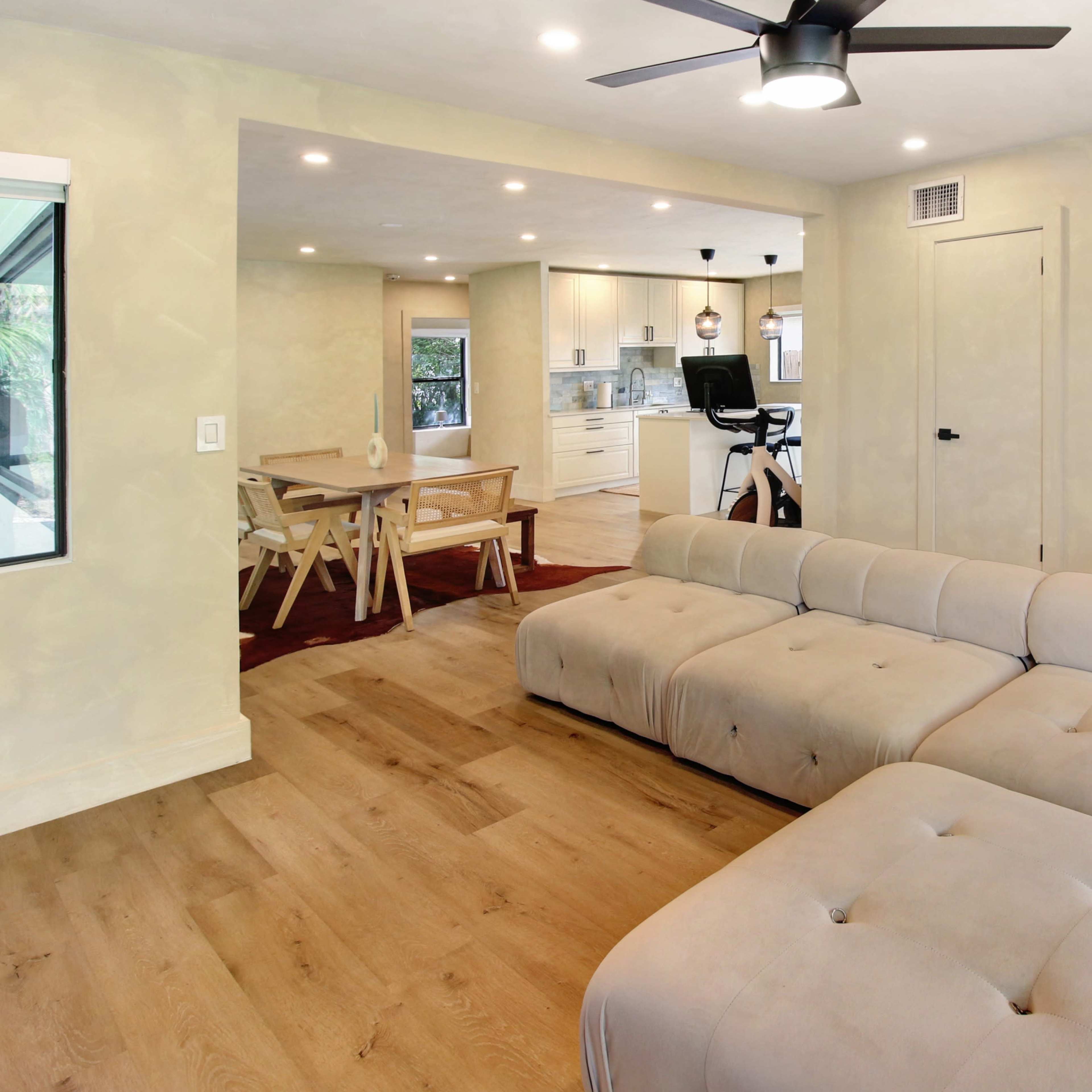A modern living room featuring a large beige sectional sofa, a wooden dining table, and an open kitchen area with bright lighting.