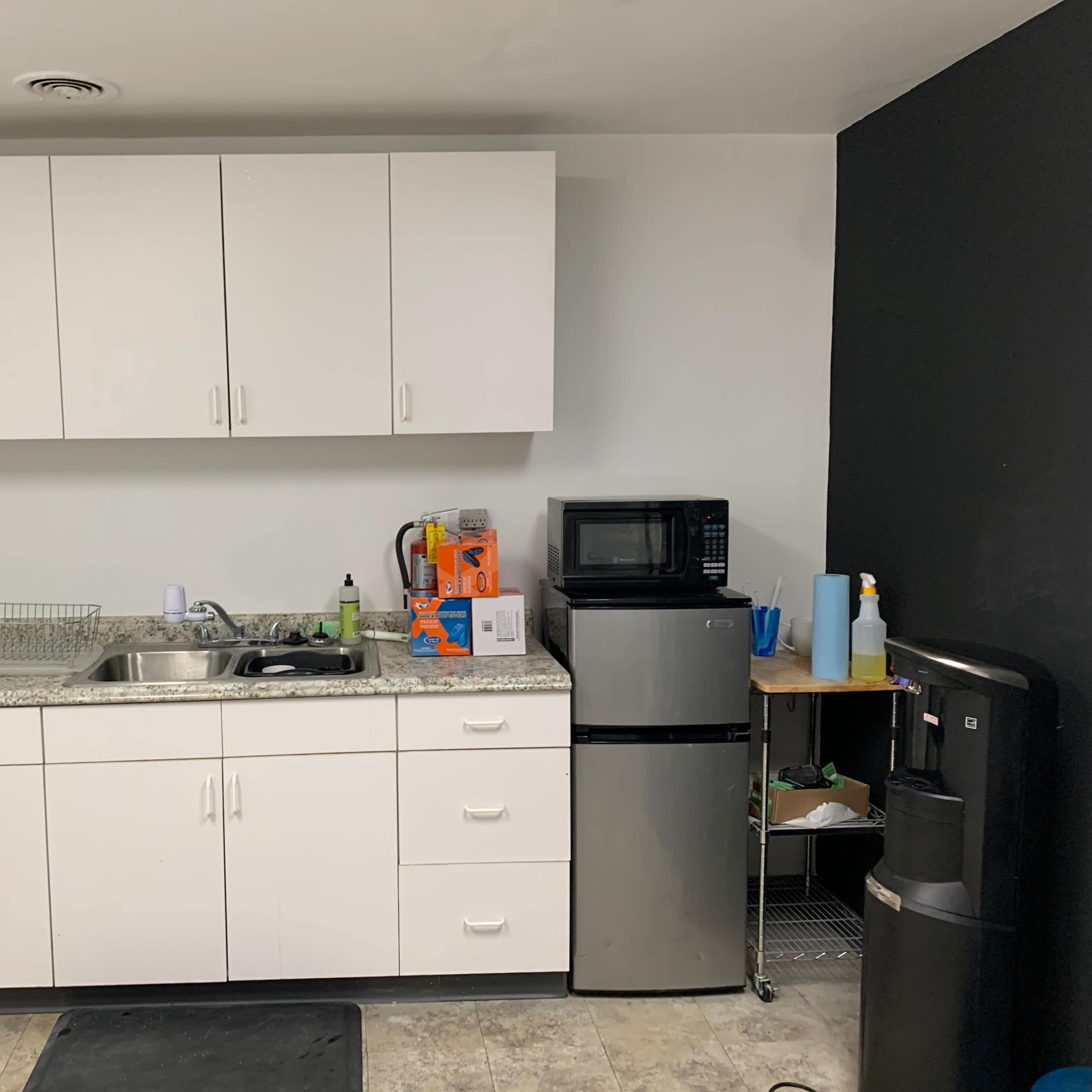 A kitchenette with white cabinets, a sink, a microwave, a mini fridge, and a water dispenser against a partially black wall.