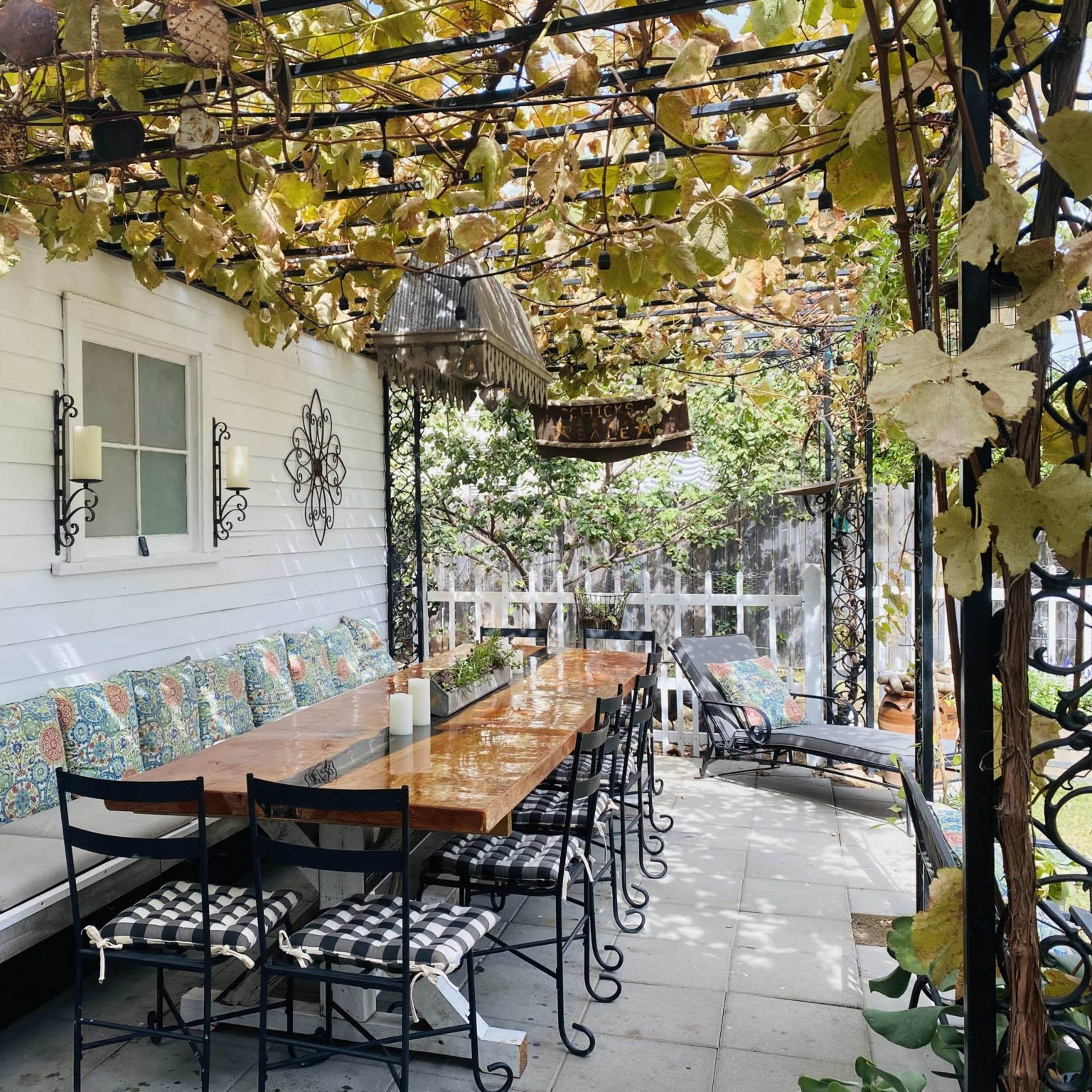 A long wooden table surrounded by black metal chairs is set under a vine-covered pergola on a patio.