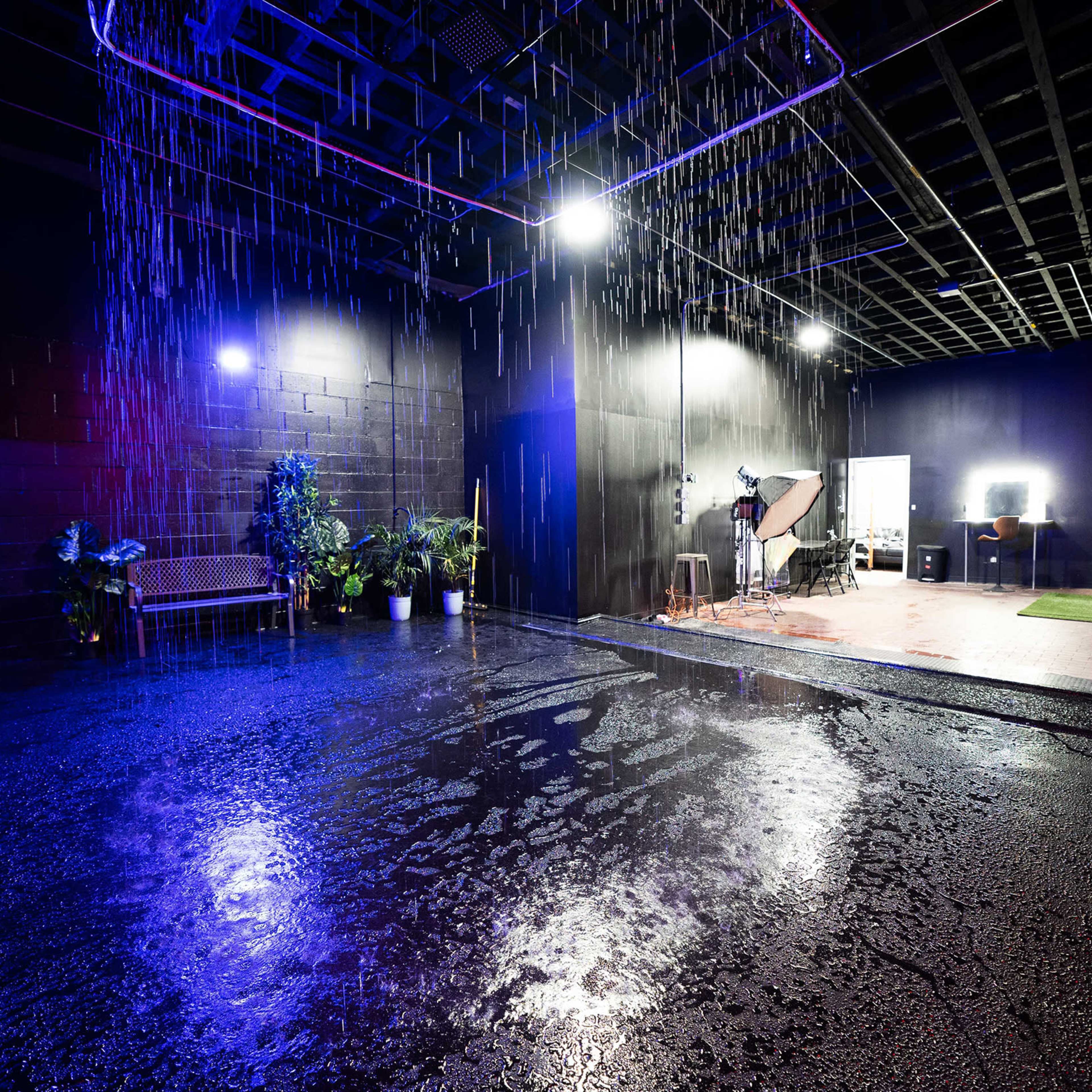 A dark indoor studio space with water falling from above, creating a wet floor and featuring various lighting setups and potted plants.