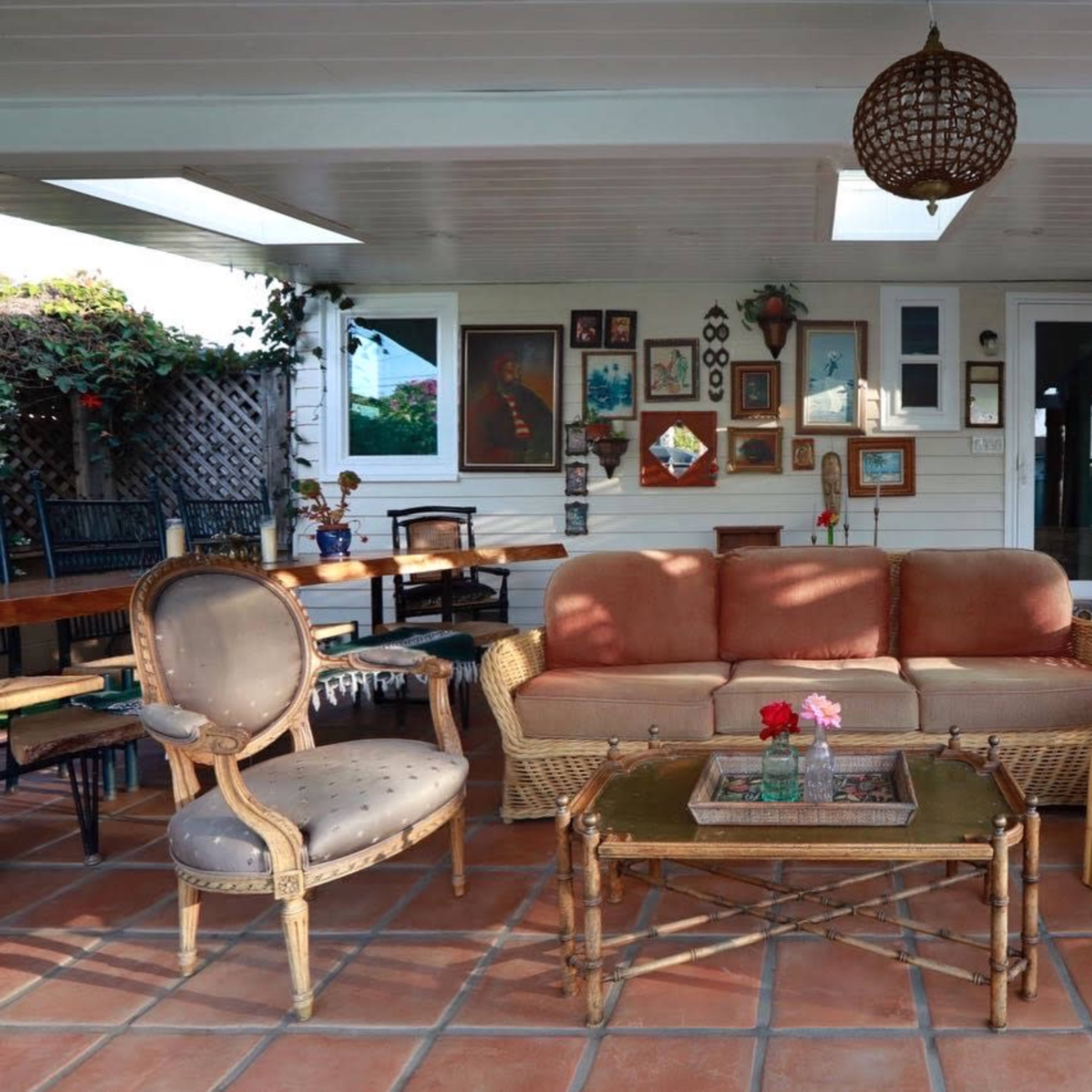 The scene features a covered outdoor seating area with a couch, two chairs, and a table on a tiled floor, surrounded by wall art and greenery.