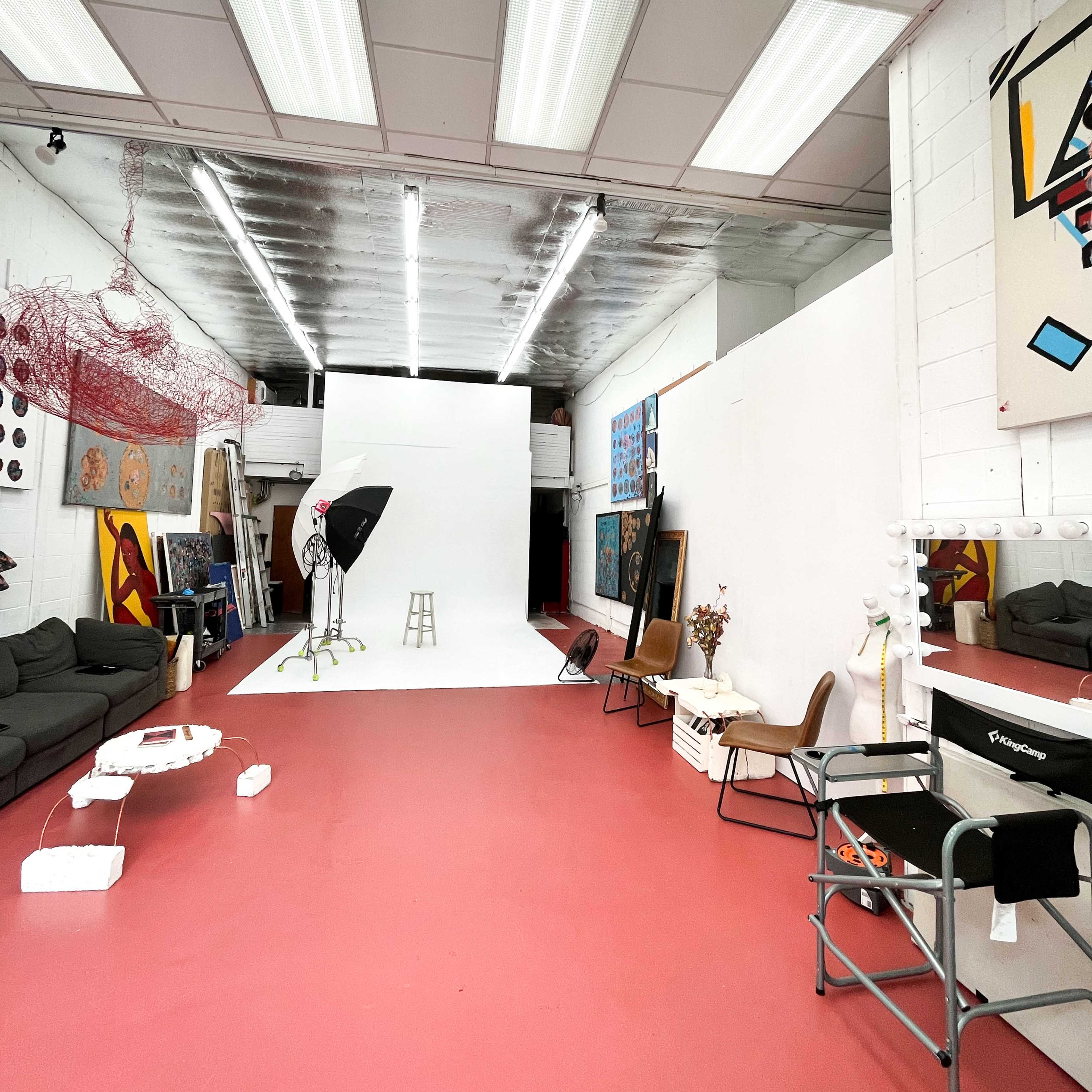A spacious photography studio features a white backdrop, various art pieces on the walls, and seating arrangements on a red floor.