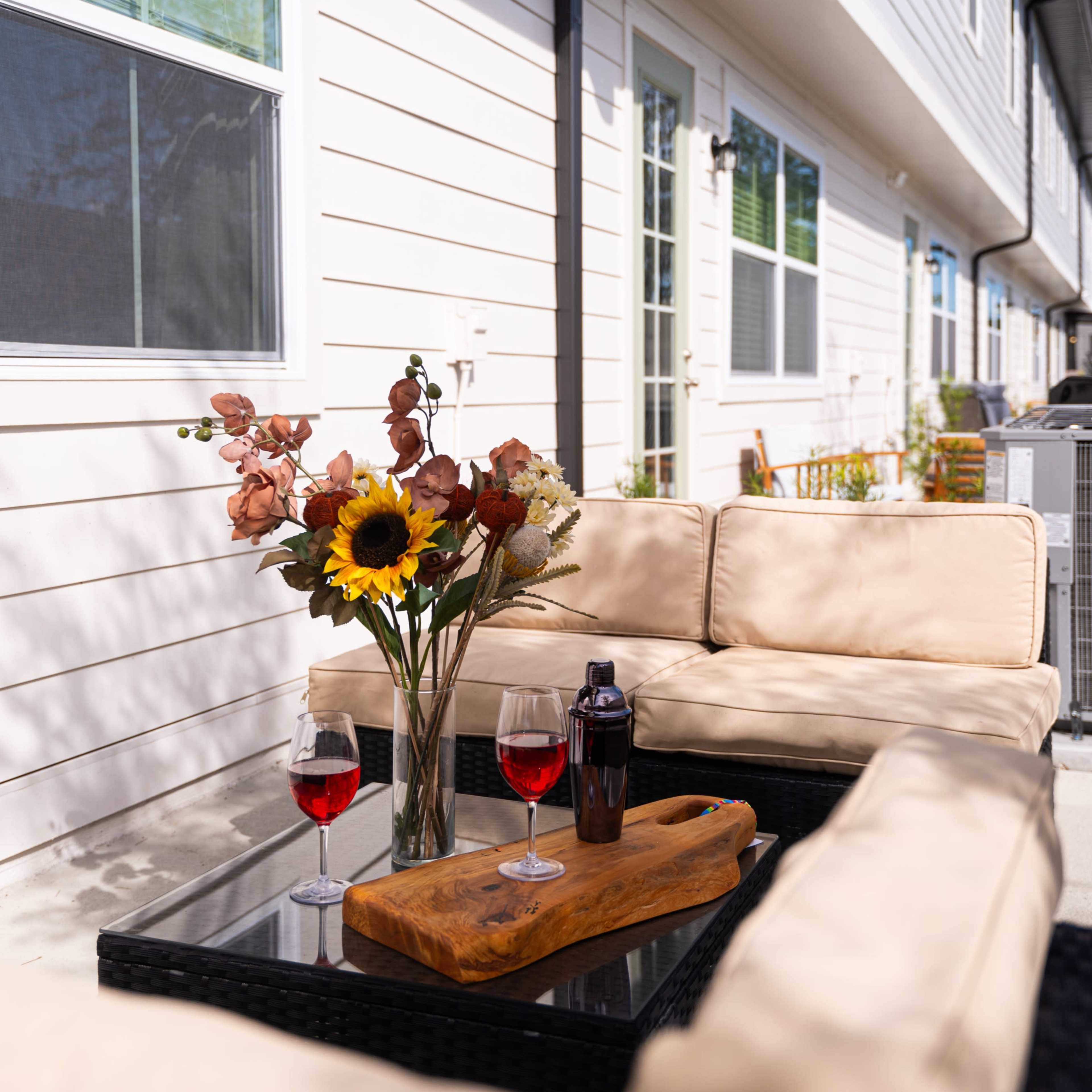 A patio seating area features a glass table with a wooden serving tray, two glasses of red drink, and a vase of flowers, located beside a white building.