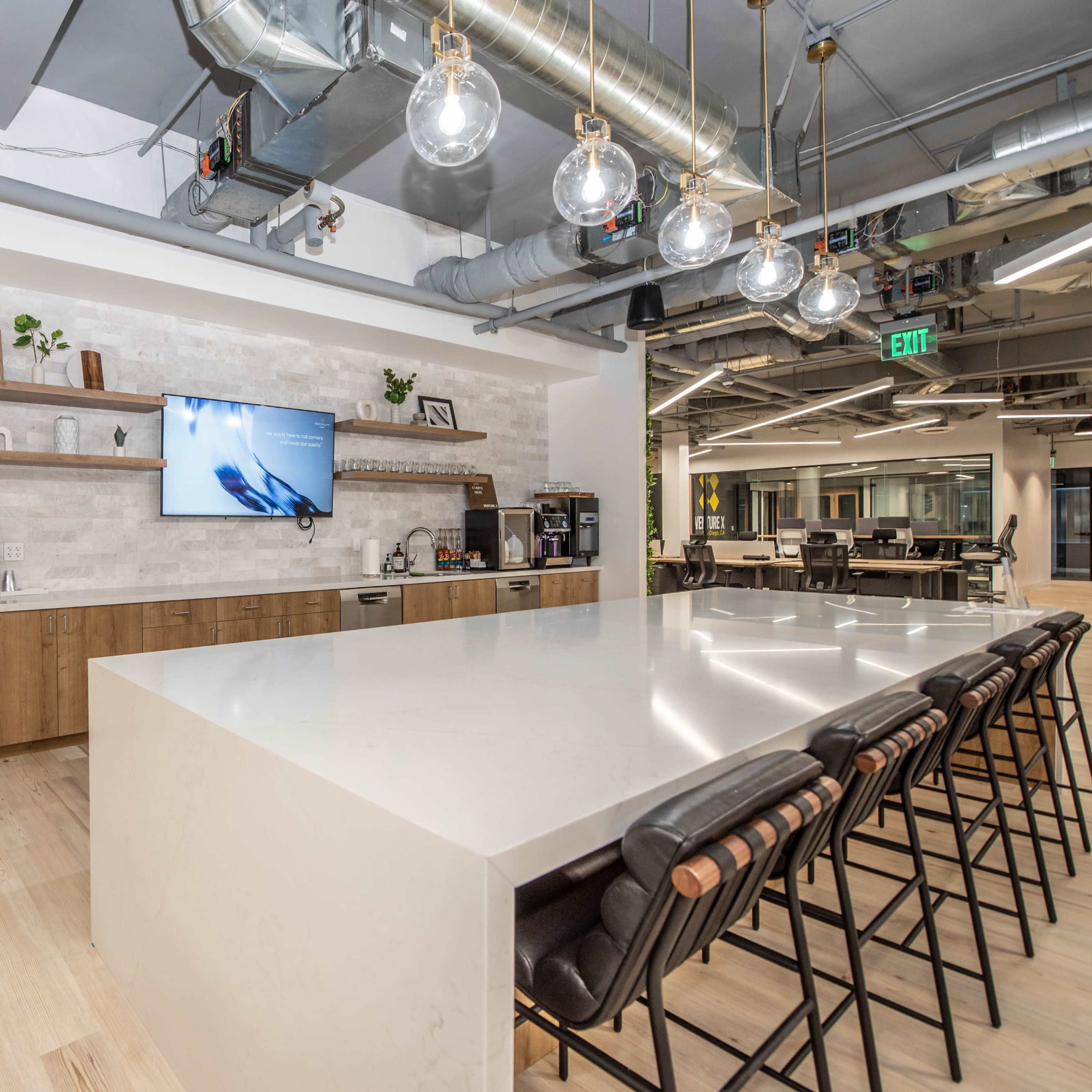 A modern office kitchen area featuring a large white countertop, wooden cabinetry, a microwave, and a television mounted on the wall.