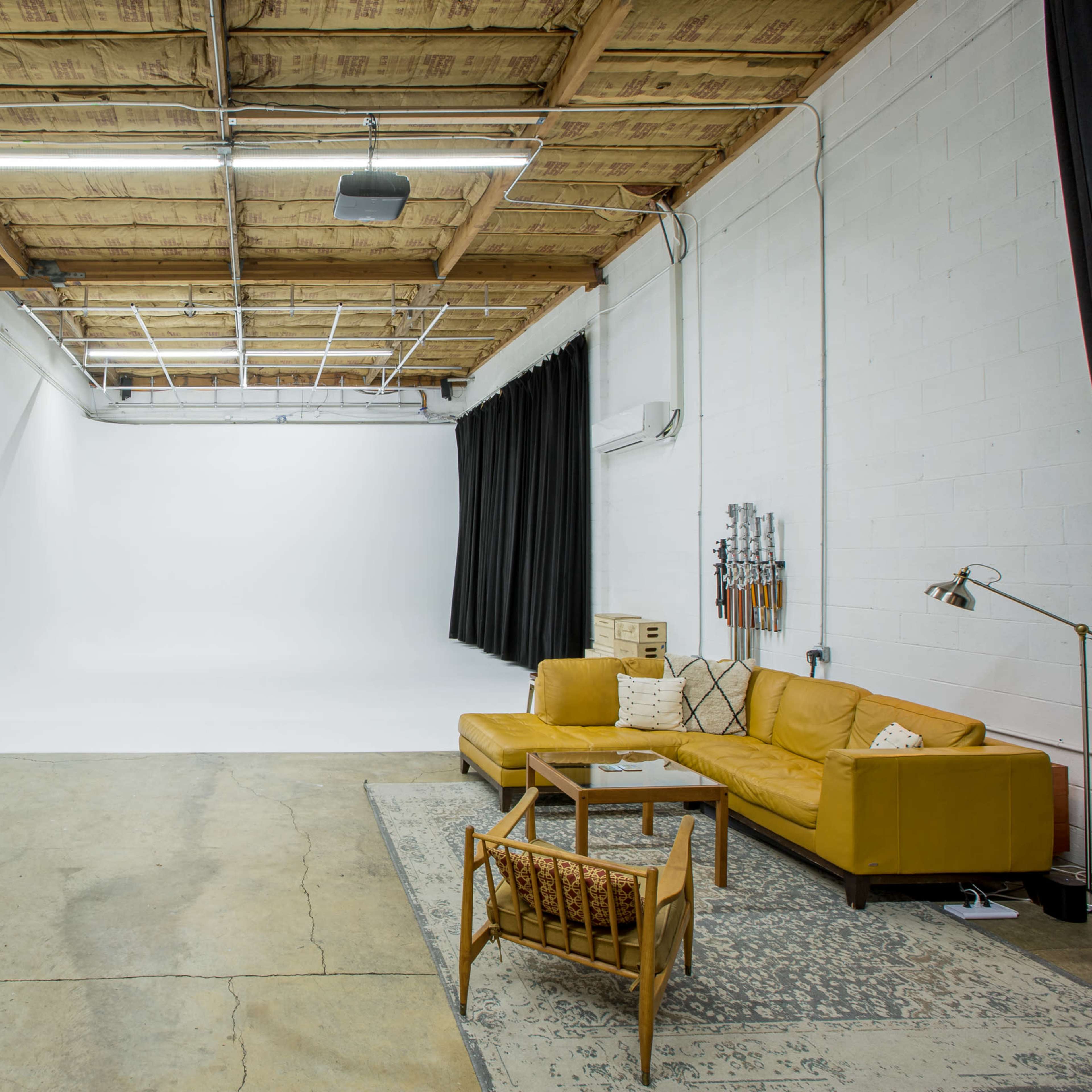 A spacious studio features a yellow sectional sofa, a wooden chair, two makeup stations with mirrors, and a backdrop of white and black curtains.