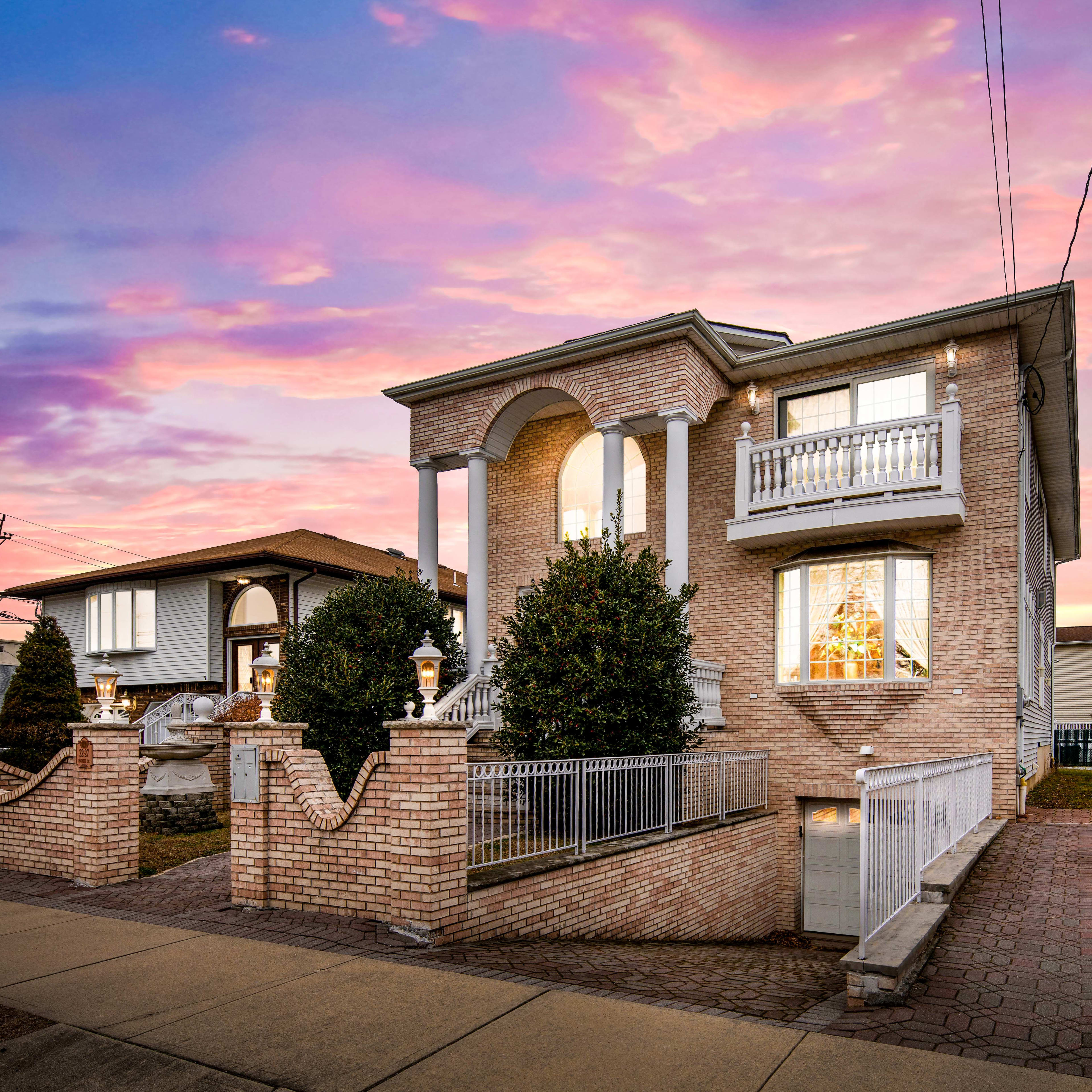 A two-story brick house with a decorative front entrance and landscaped yard is illuminated by a colorful sunset.