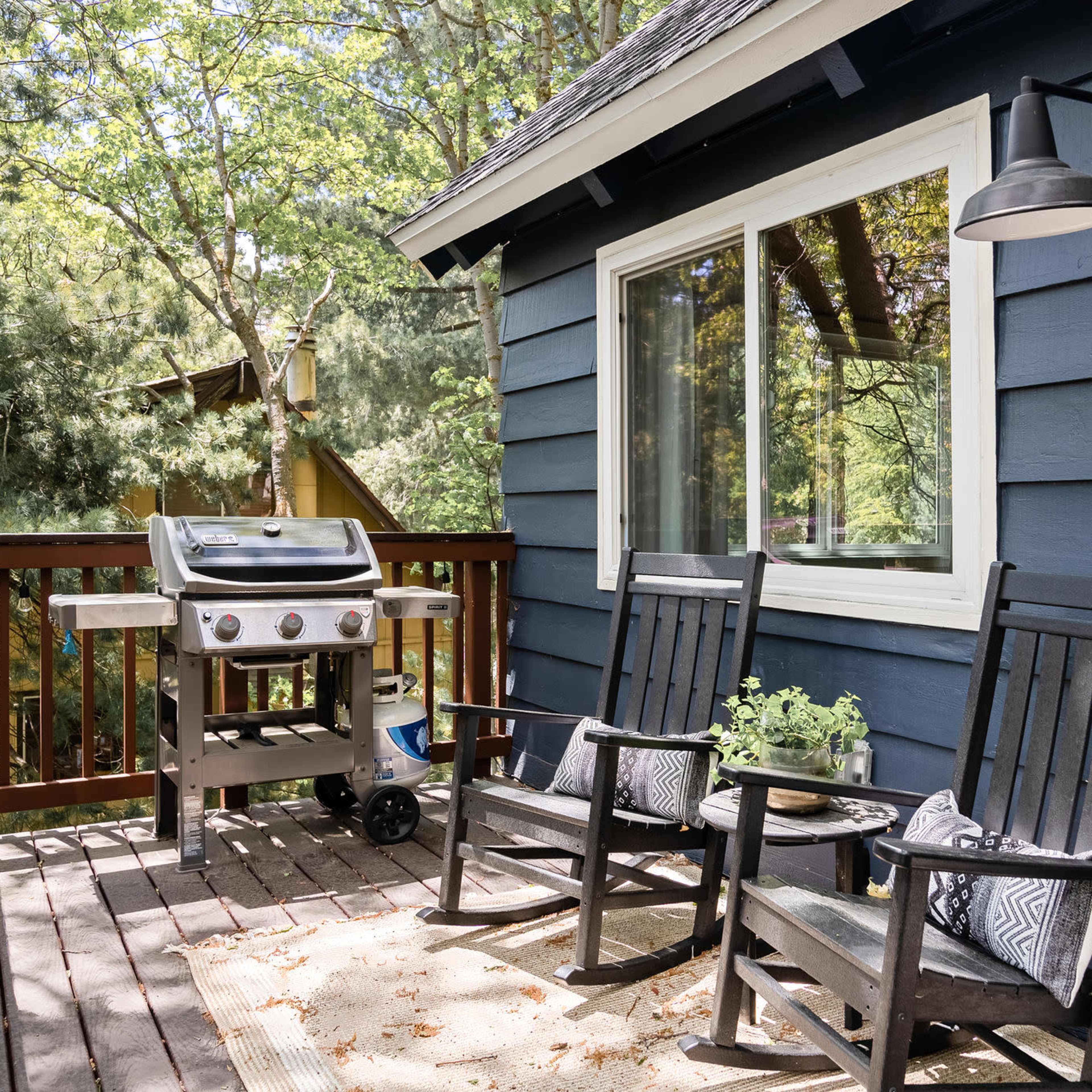 A wooden deck features two rocking chairs and a gas grill beside a large window, surrounded by greenery.