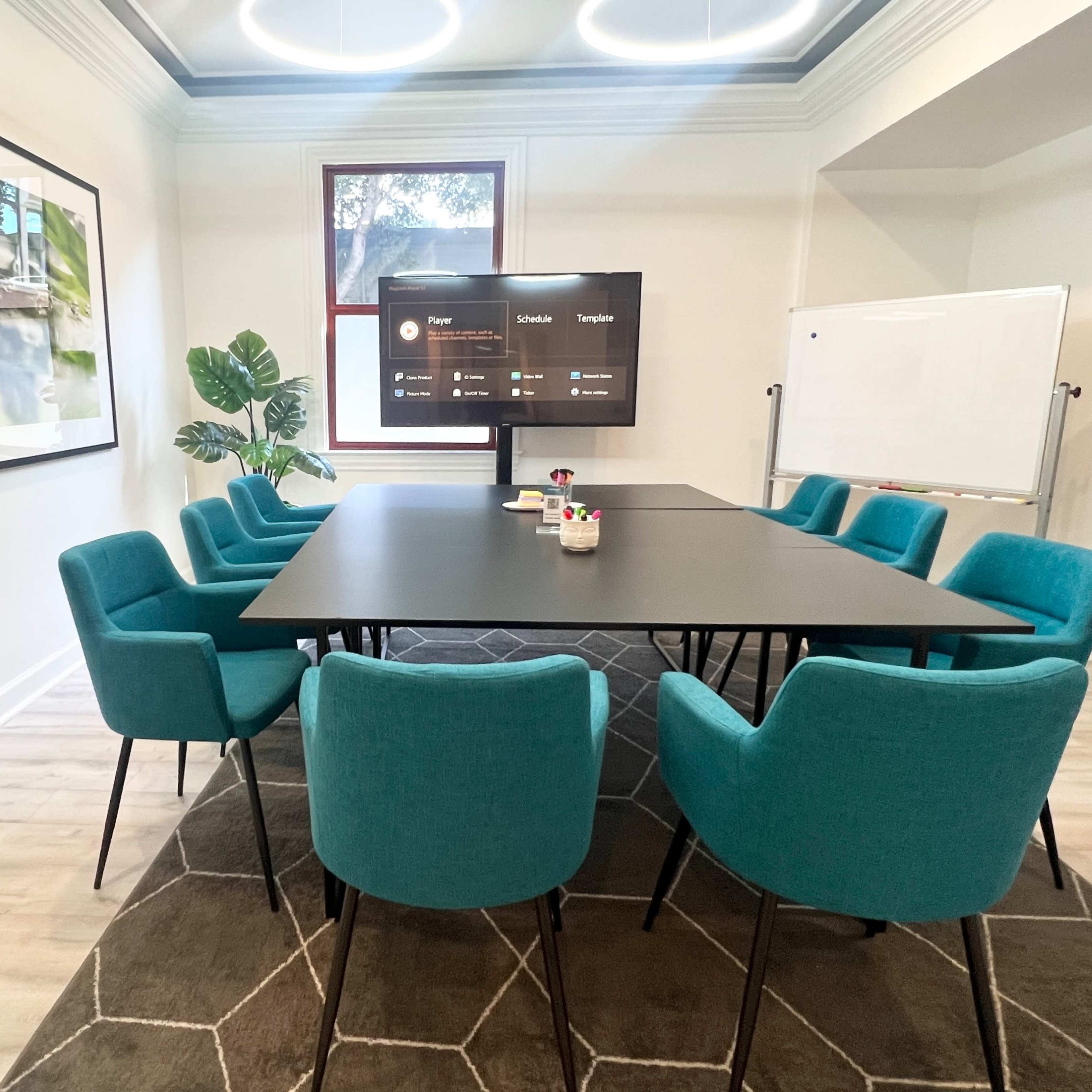 A meeting room features a large black table surrounded by teal chairs, a presentation screen, and framed nature photographs on the walls.