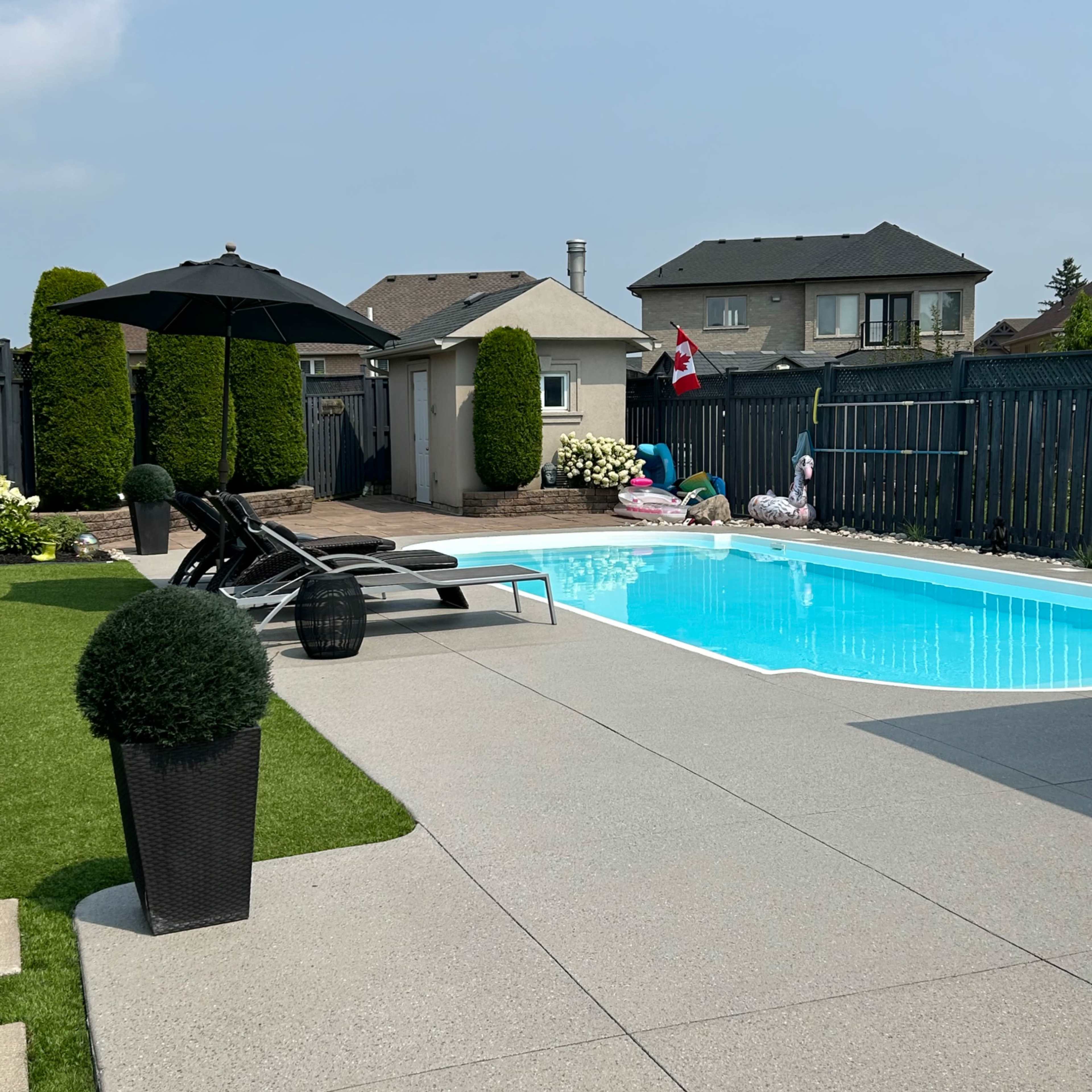 A backyard with a swimming pool, lounge chairs, and a patio area surrounded by fencing and greenery.