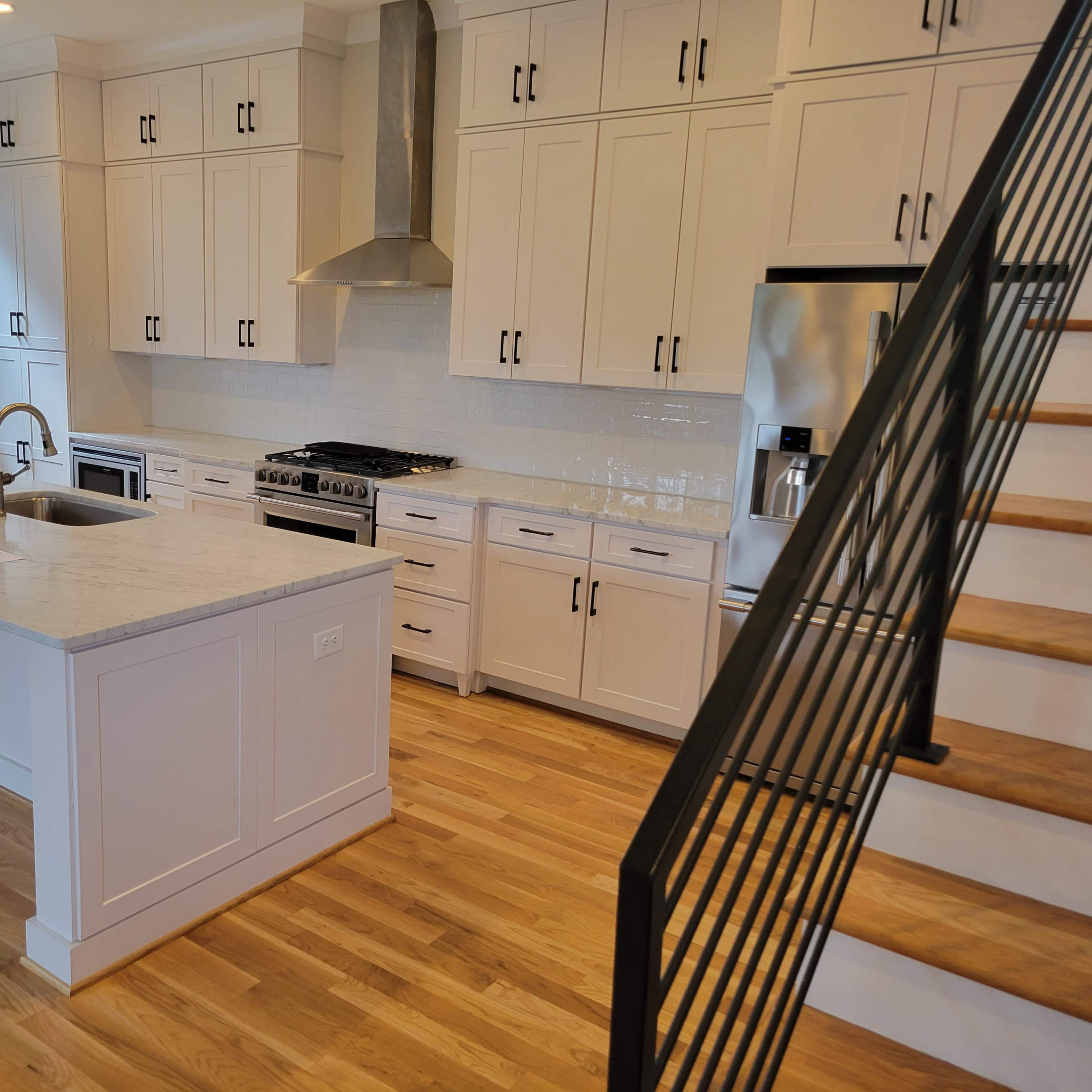 A modern kitchen with white cabinetry, a large island, stainless steel appliances, and a wooden staircase in the background.