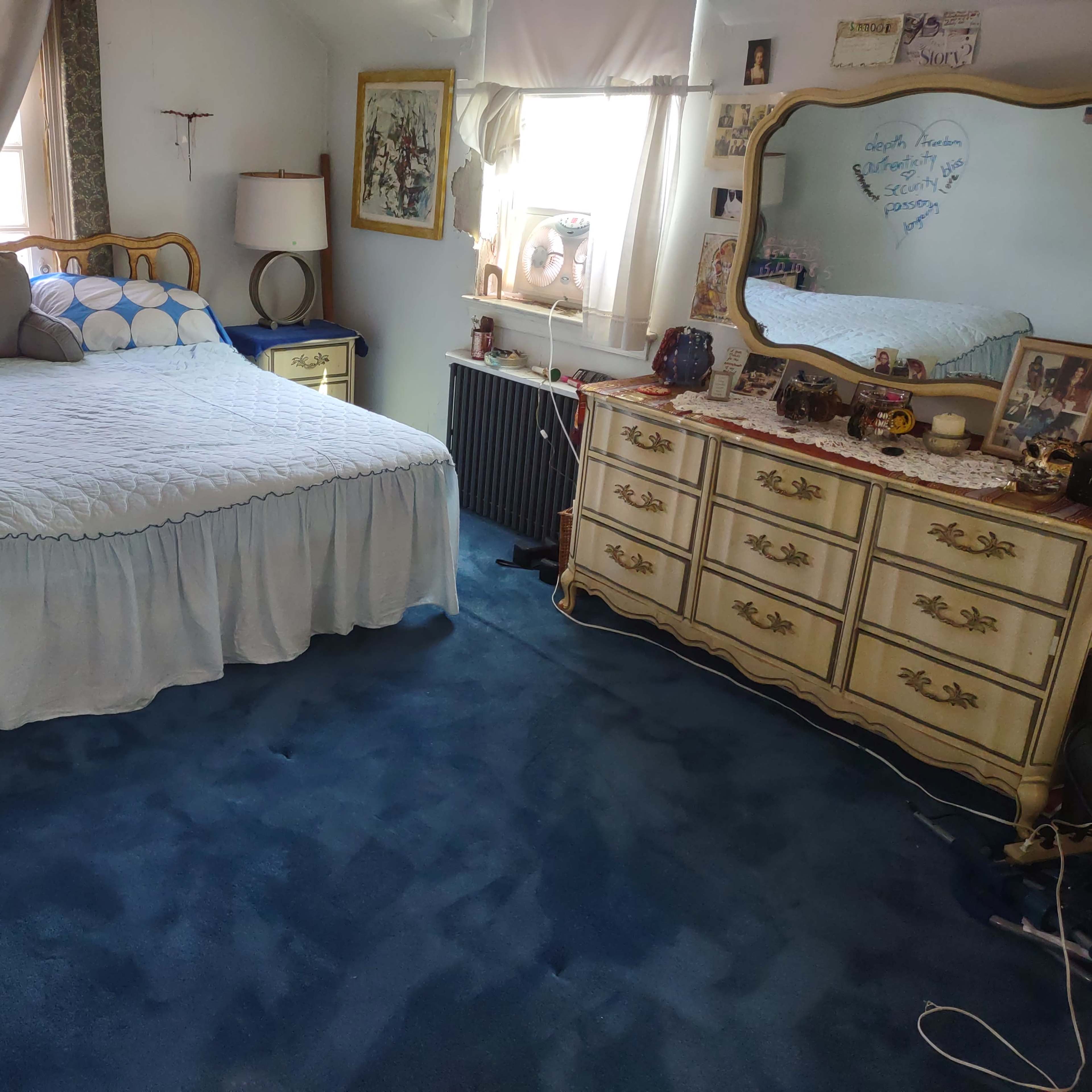 The room features a bed with a blue and white bedspread, a wooden dresser, and a large mirror, all situated on a blue carpet.
