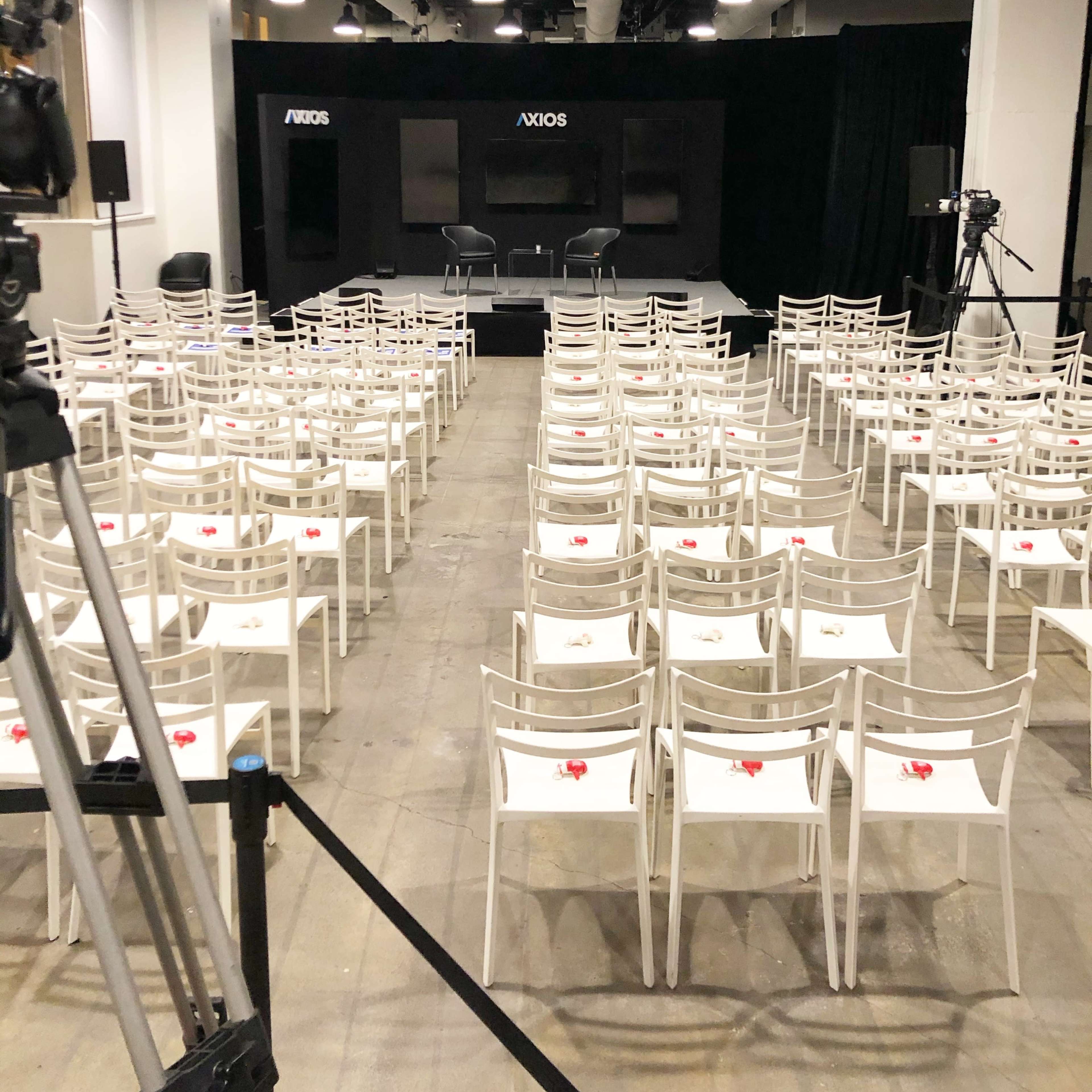 A spacious indoor venue is set up for an event, featuring rows of empty white chairs facing a stage with Axios branding and cameras positioned for recording.