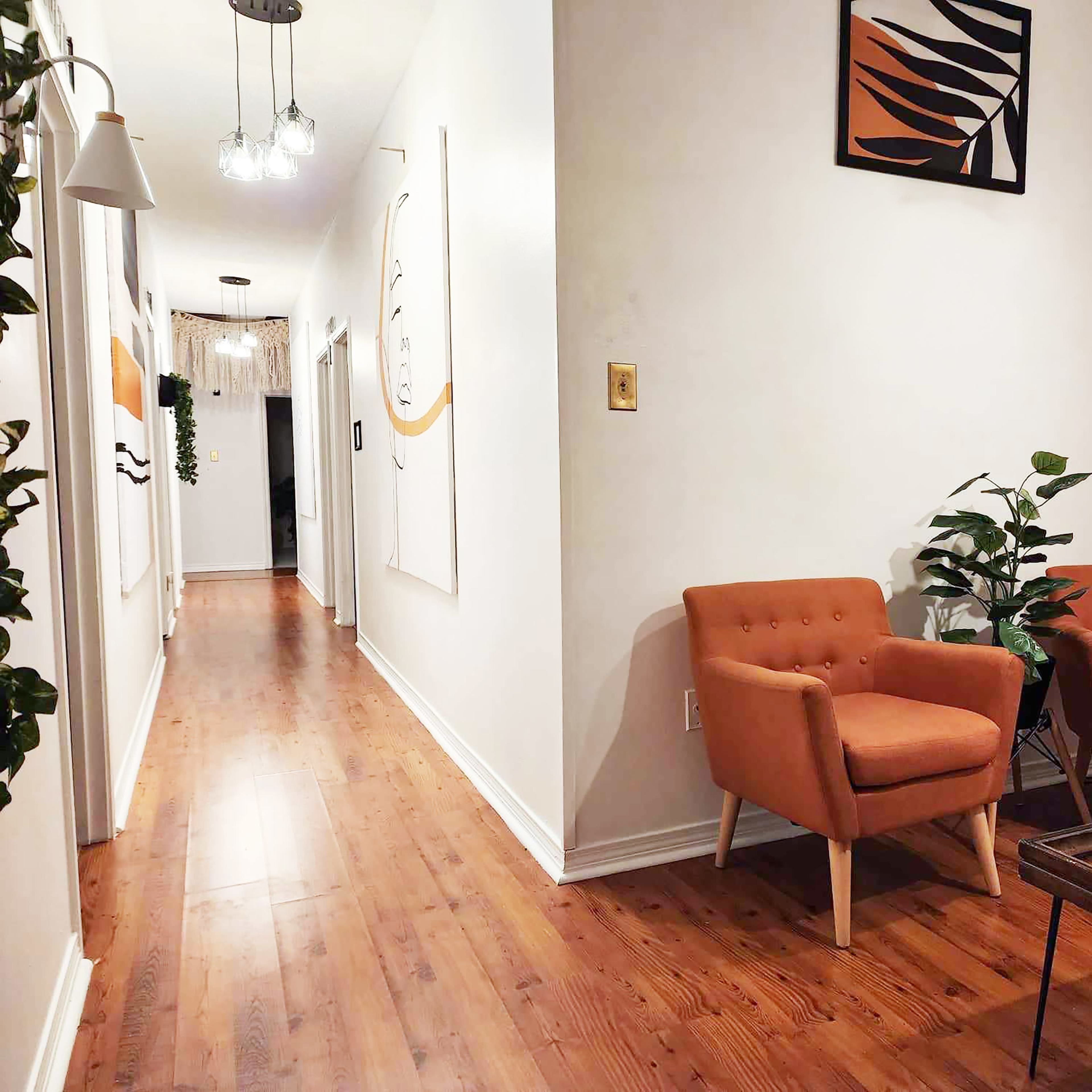 A narrow hallway leading to a living area with two orange armchairs, a coffee table, and decorative wall art.