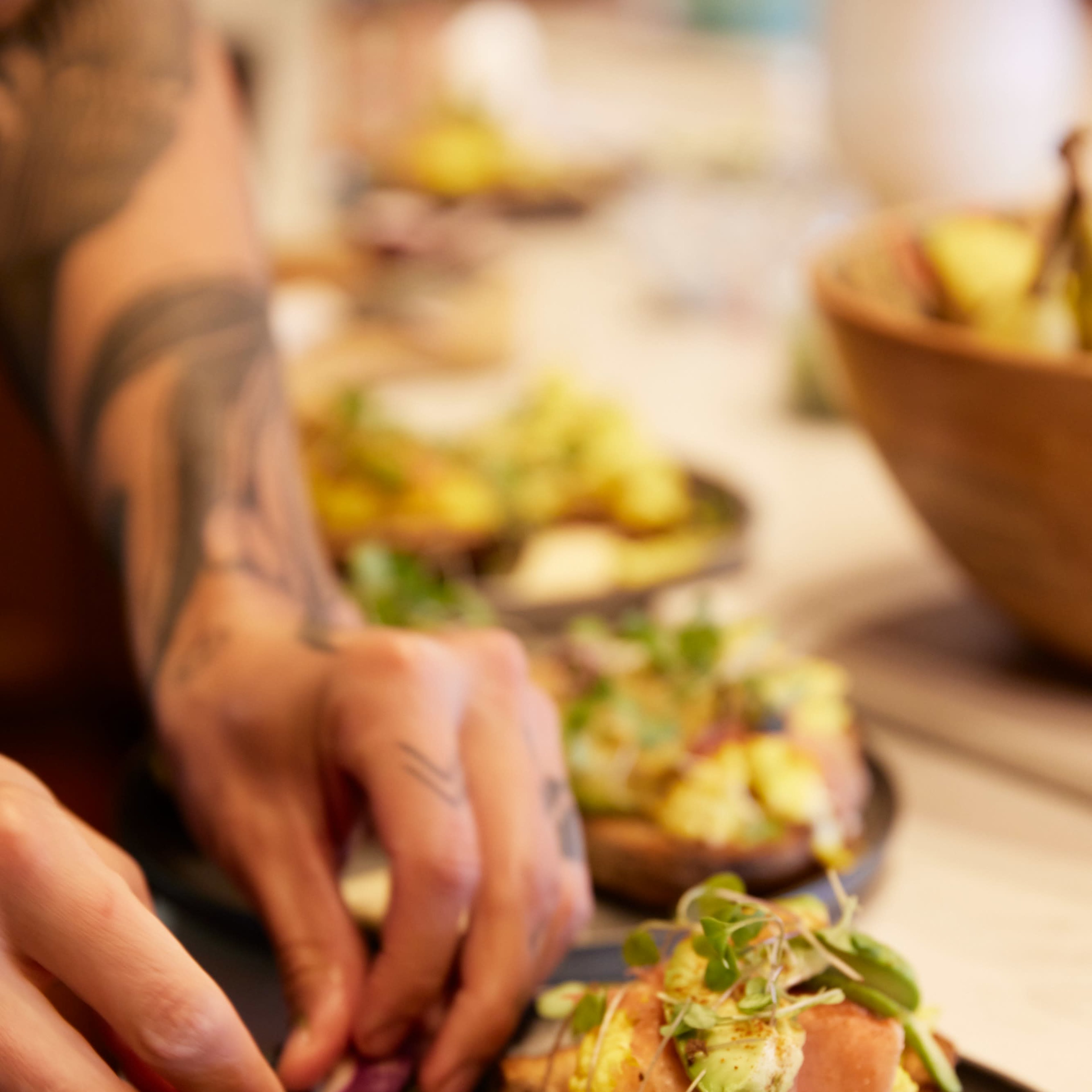 A person is arranging thinly sliced ham and microgreens on toasted bread topped with scrambled eggs on a dark plate.