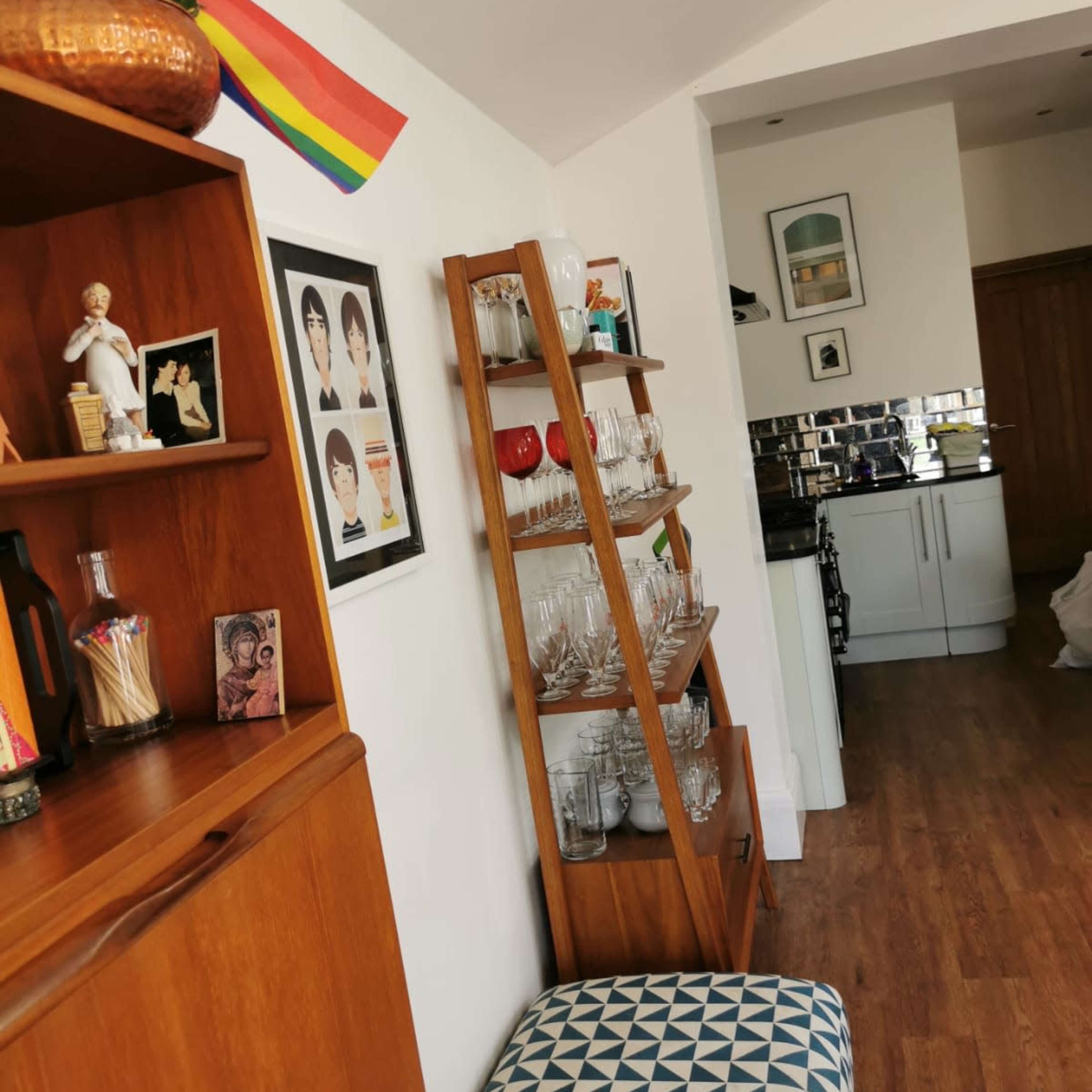 A living space featuring a wooden shelf with glassware, a decorated wall with framed art, and a glimpse of a kitchen in the background.