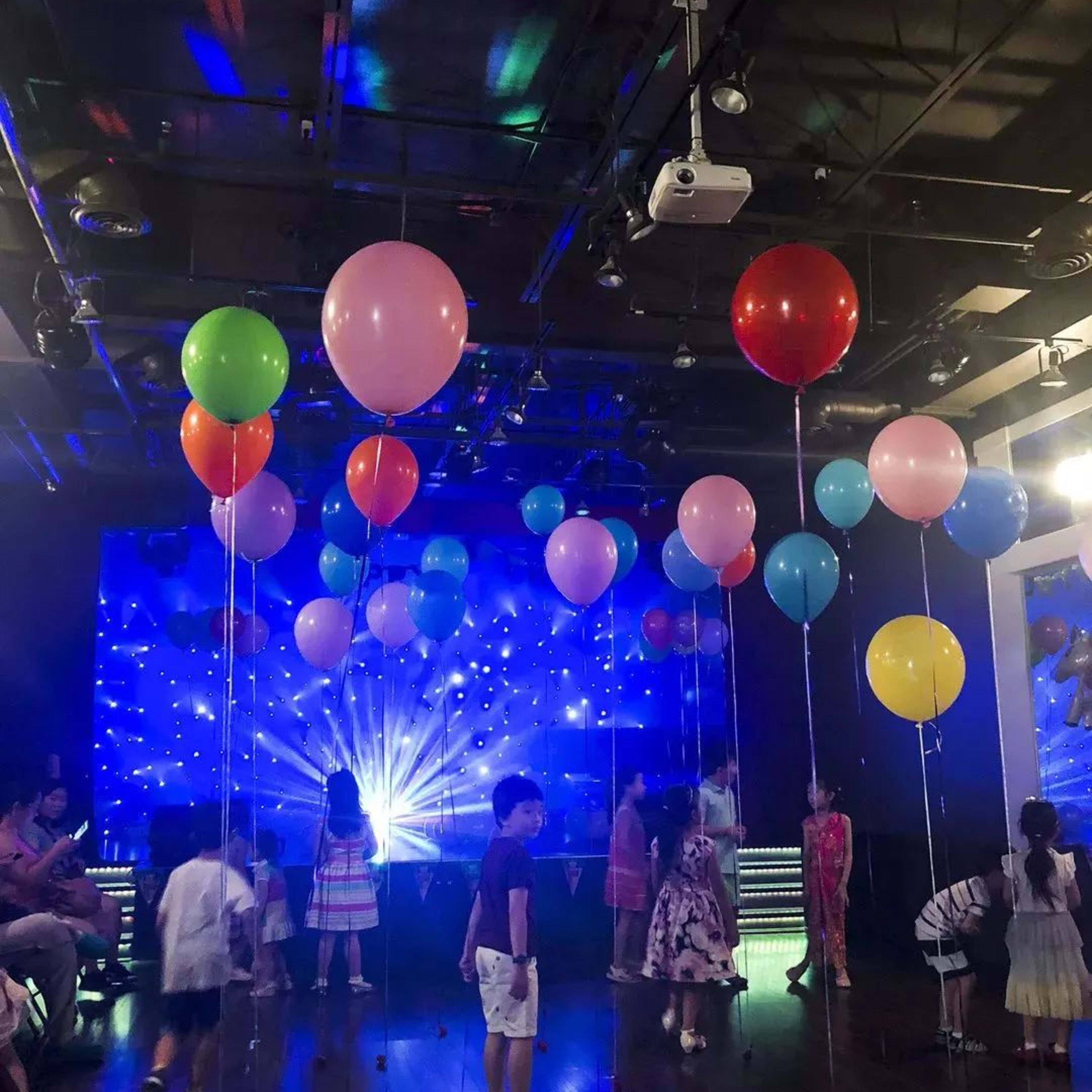 Children are playing with colorful balloons in a brightly lit party room with a light display.