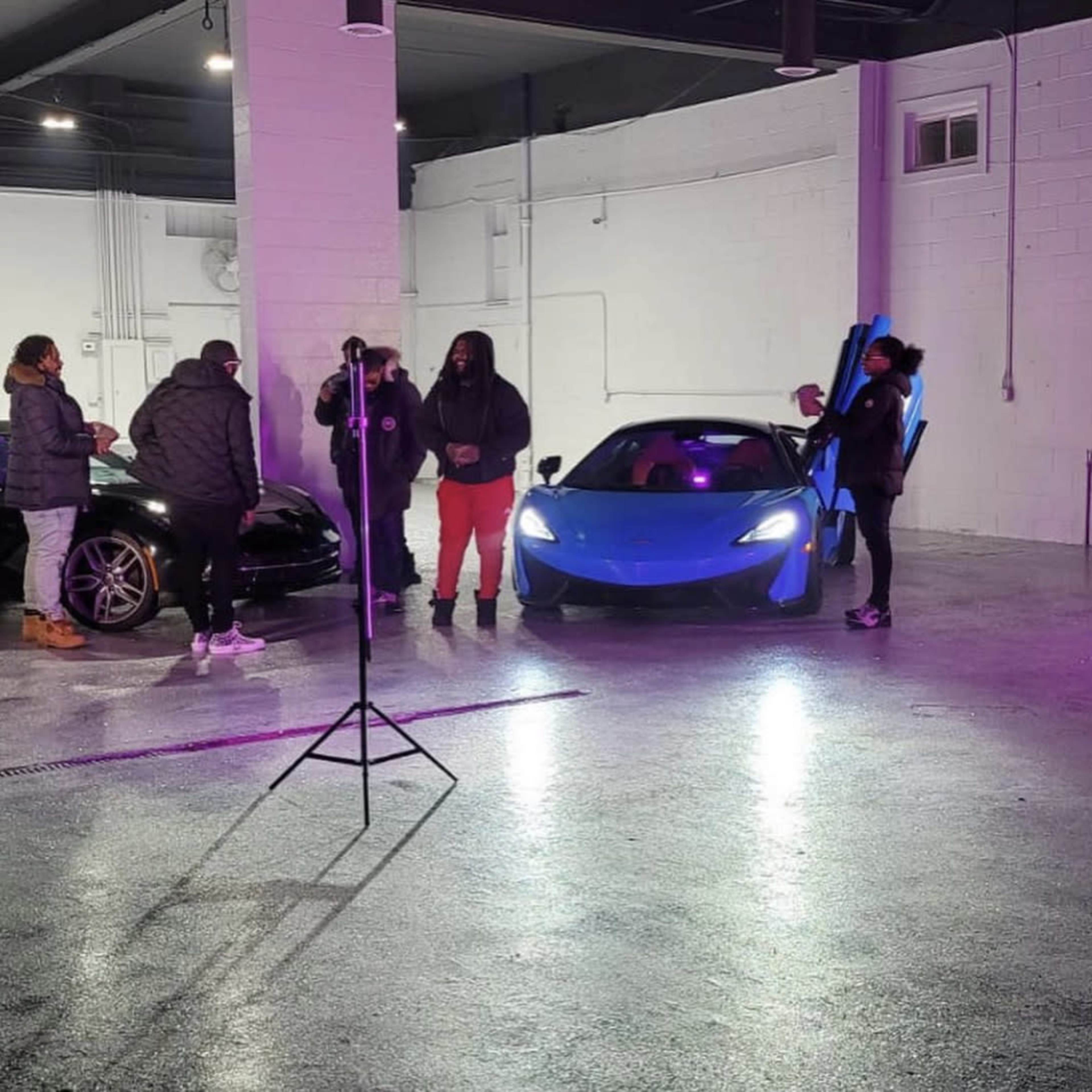 A group of people is gathered around a blue sports car in a brightly lit garage, with another black car parked nearby.
