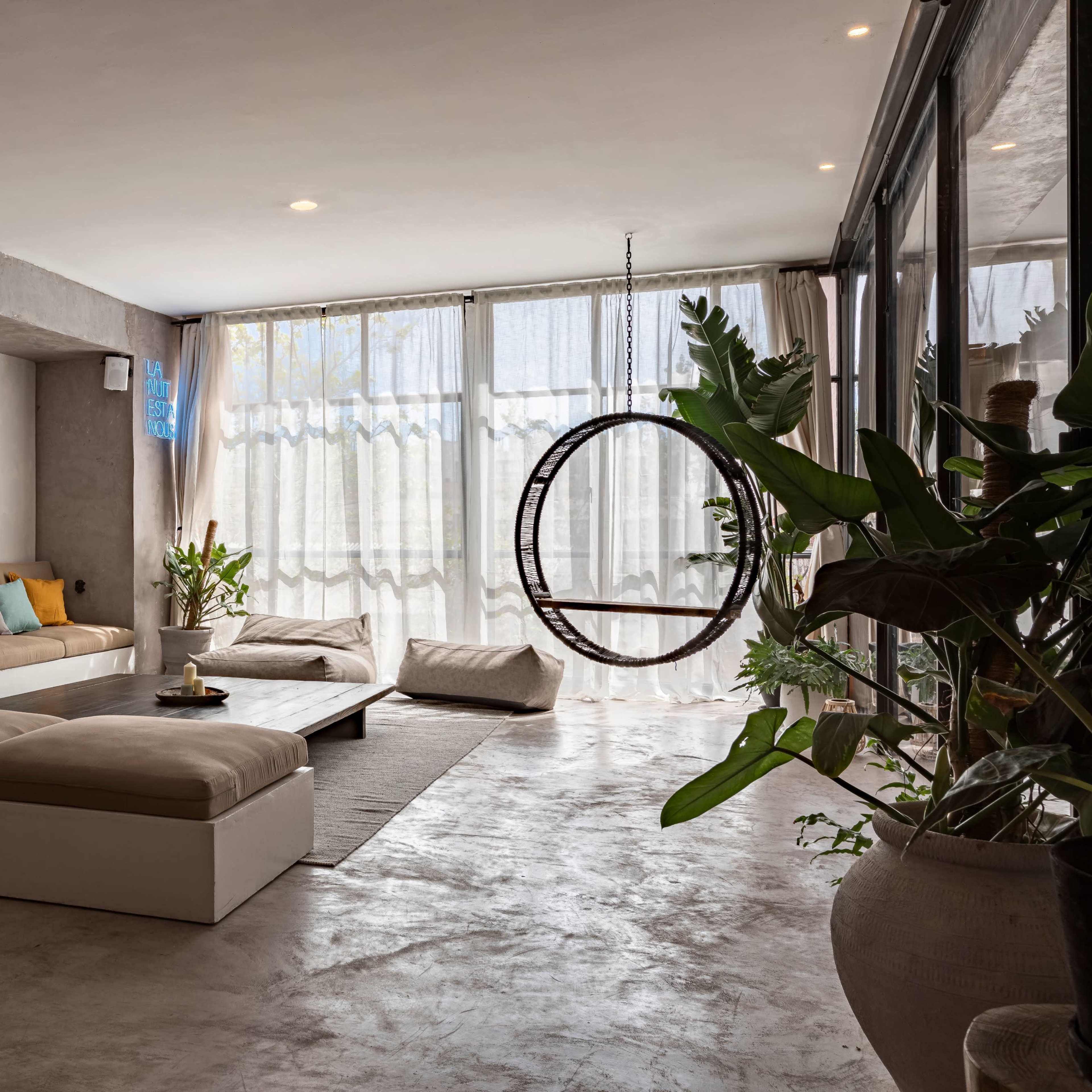 A spacious living room features a large beige sectional sofa with colorful throw pillows, large windows with sheer curtains, and a circular hanging chair.