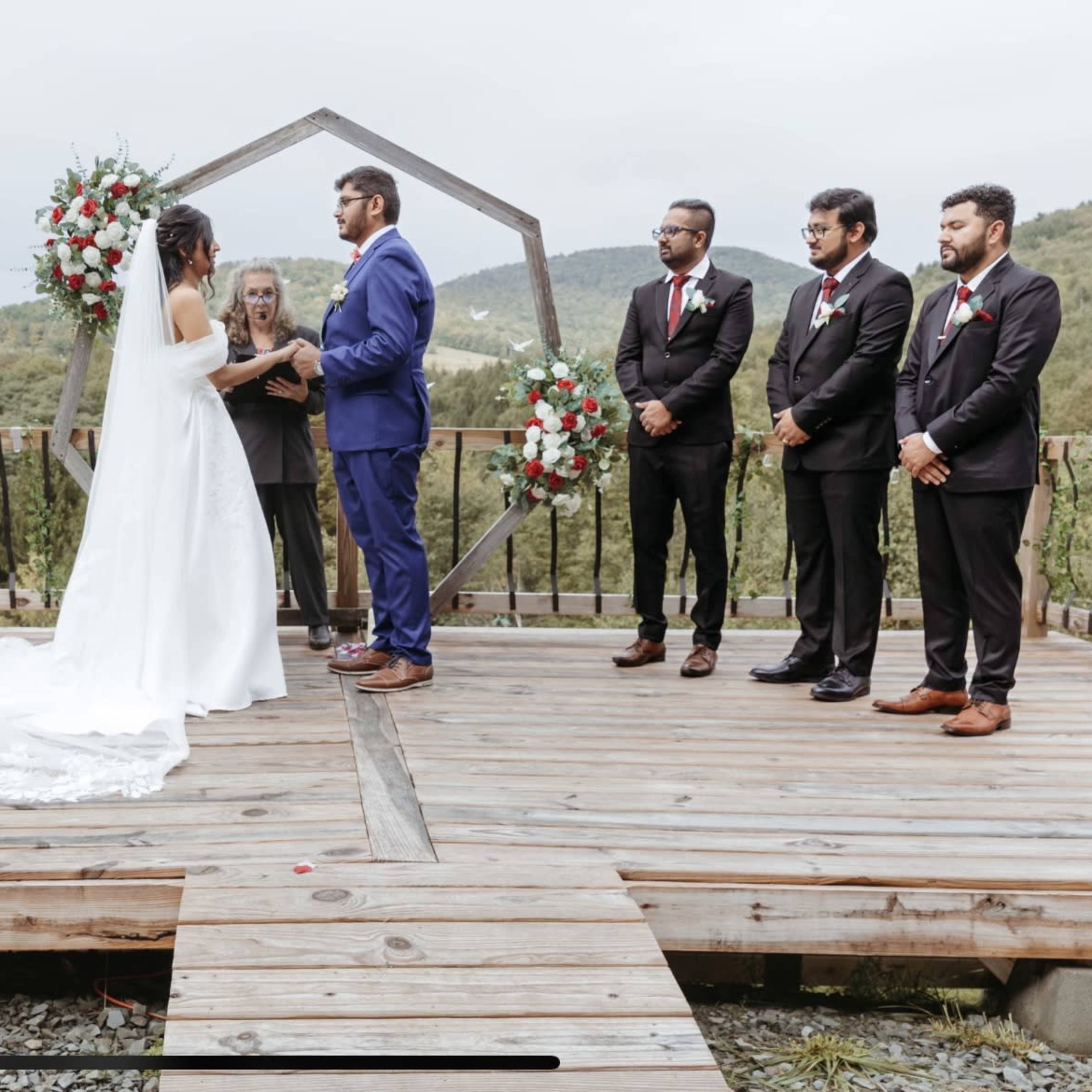 A couple exchanges vows under a decorative arch surrounded by their wedding party on a wooden platform in a scenic outdoor setting.