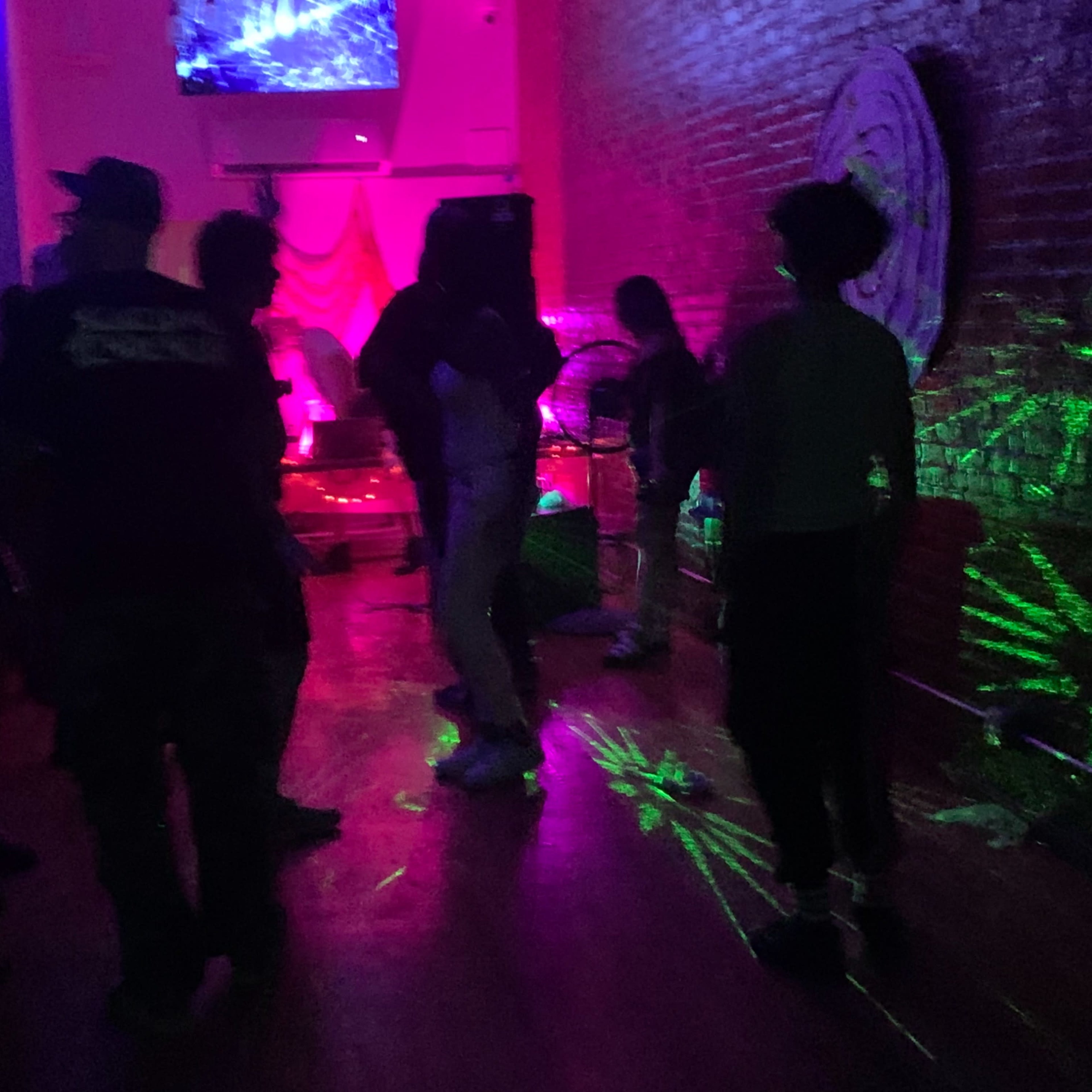 A group of people dance under colorful lights in a dimly lit venue with a stage in the background.