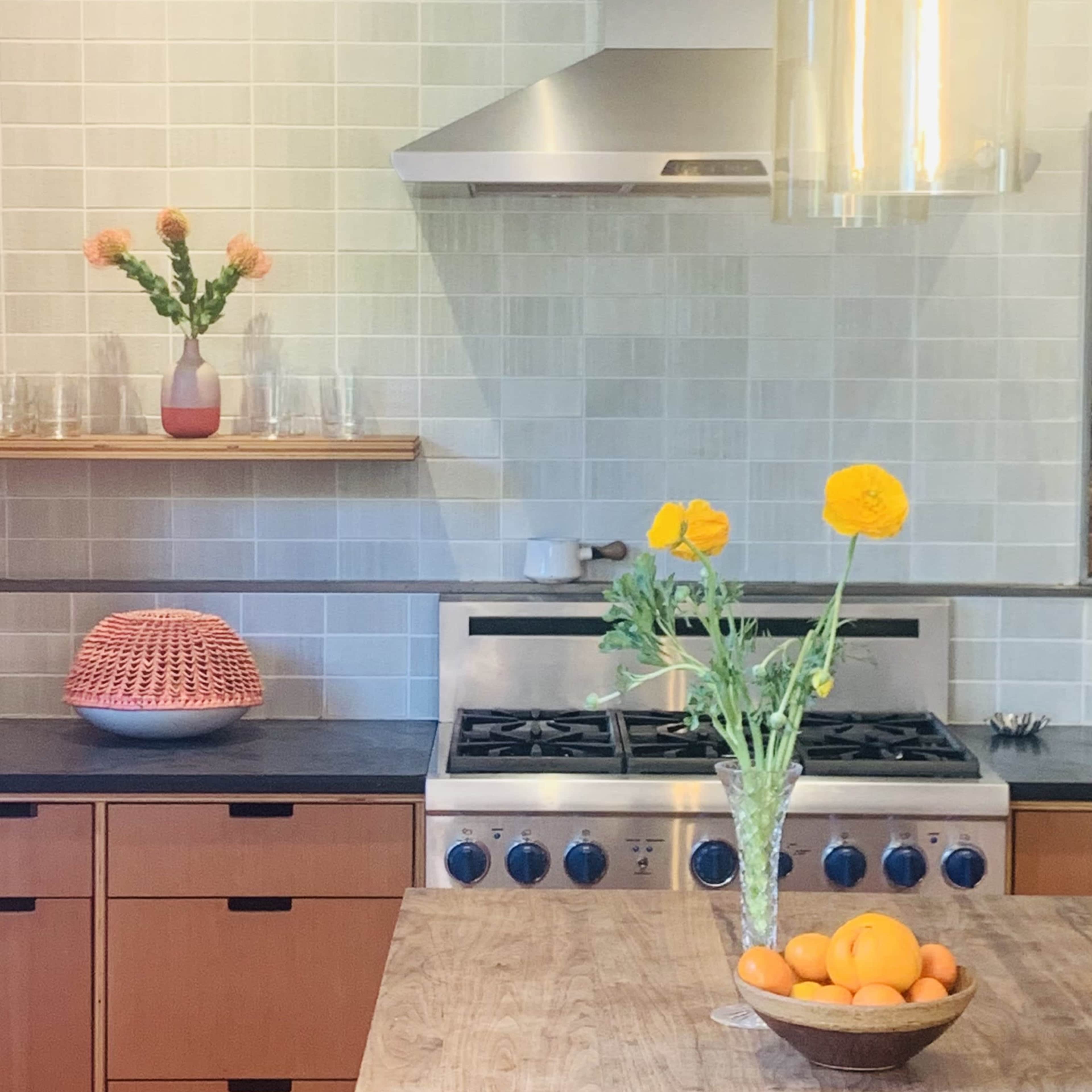 A modern kitchen features a stainless steel stove, a wooden table with a bowl of oranges, and a vase with flowers on the counter.