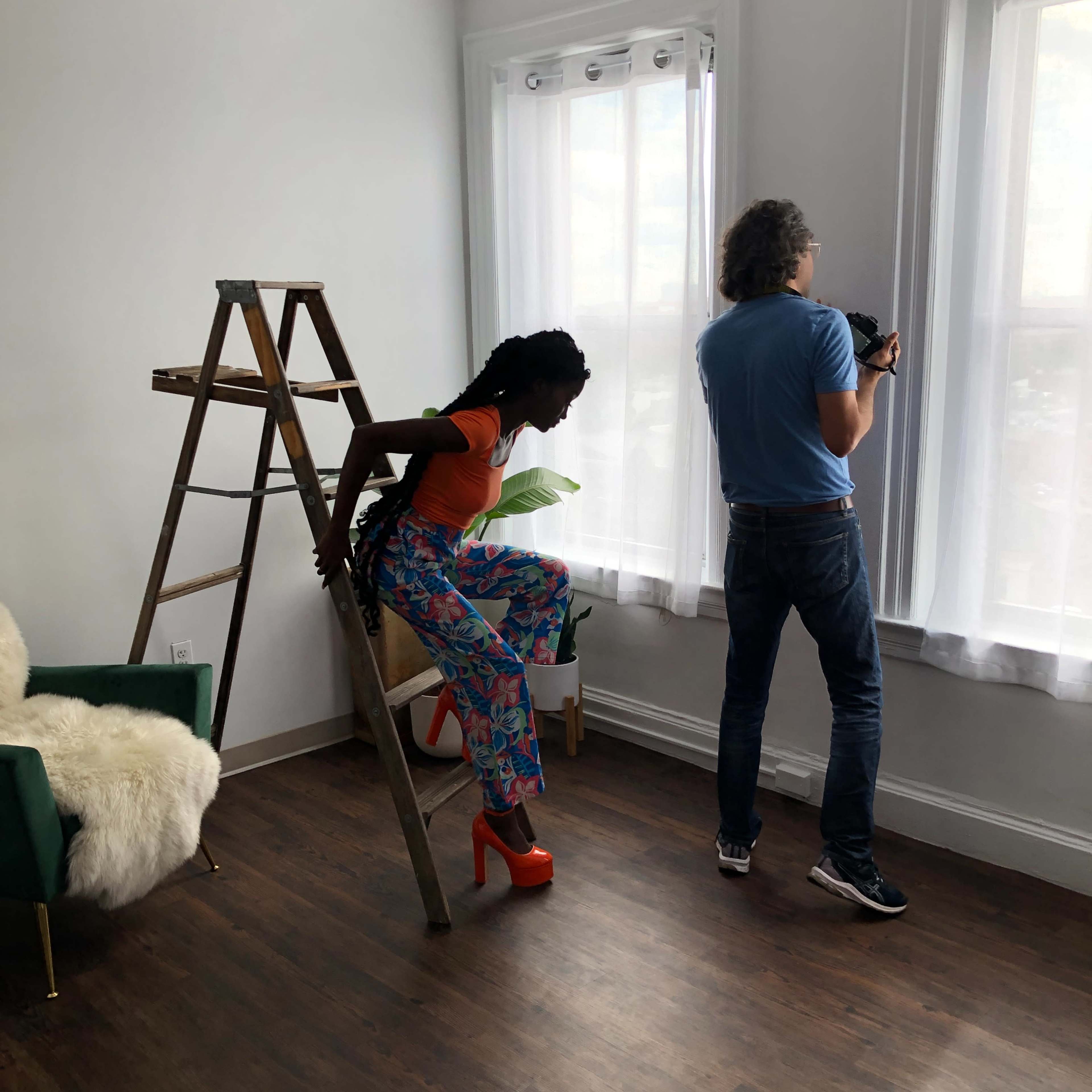 A person is sitting on a ladder while another person stands nearby, taking photos near a window in a brightly lit room.