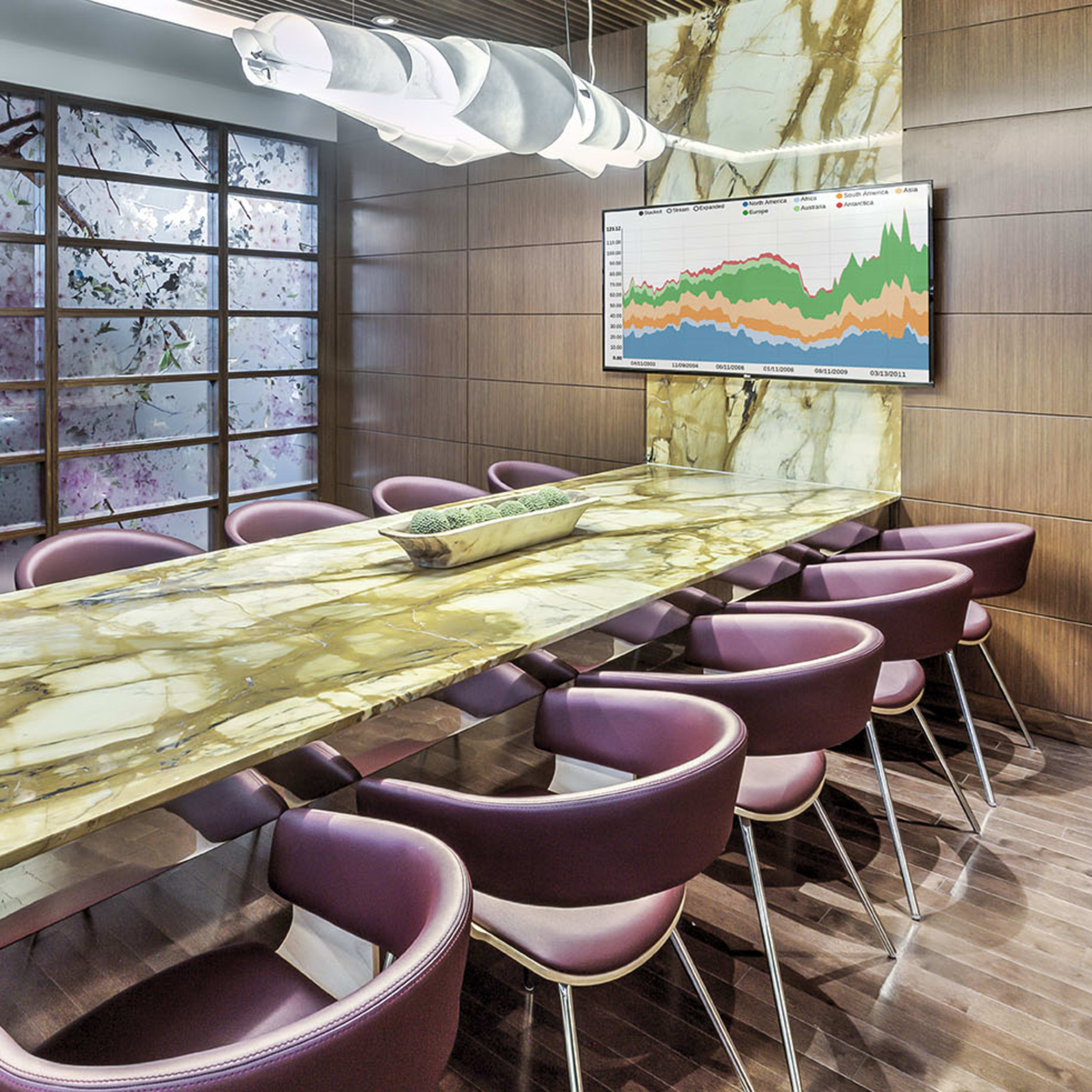 A conference room features a large marble table surrounded by purple chairs, with a wall displaying a graphic and a backdrop of frosted glass.