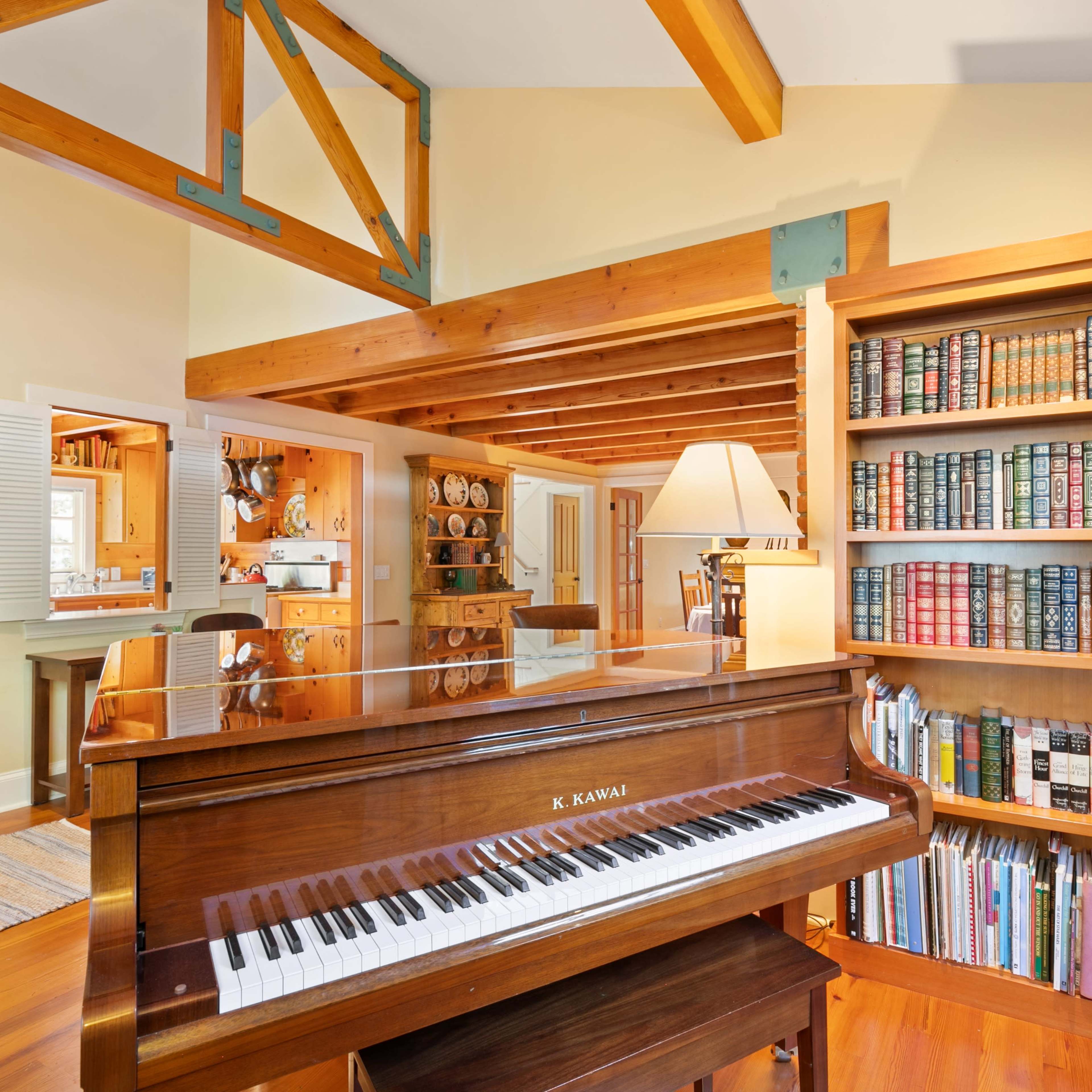 A wooden piano stands in a brightly lit room with a bookshelf filled with books and decorative items in the background.