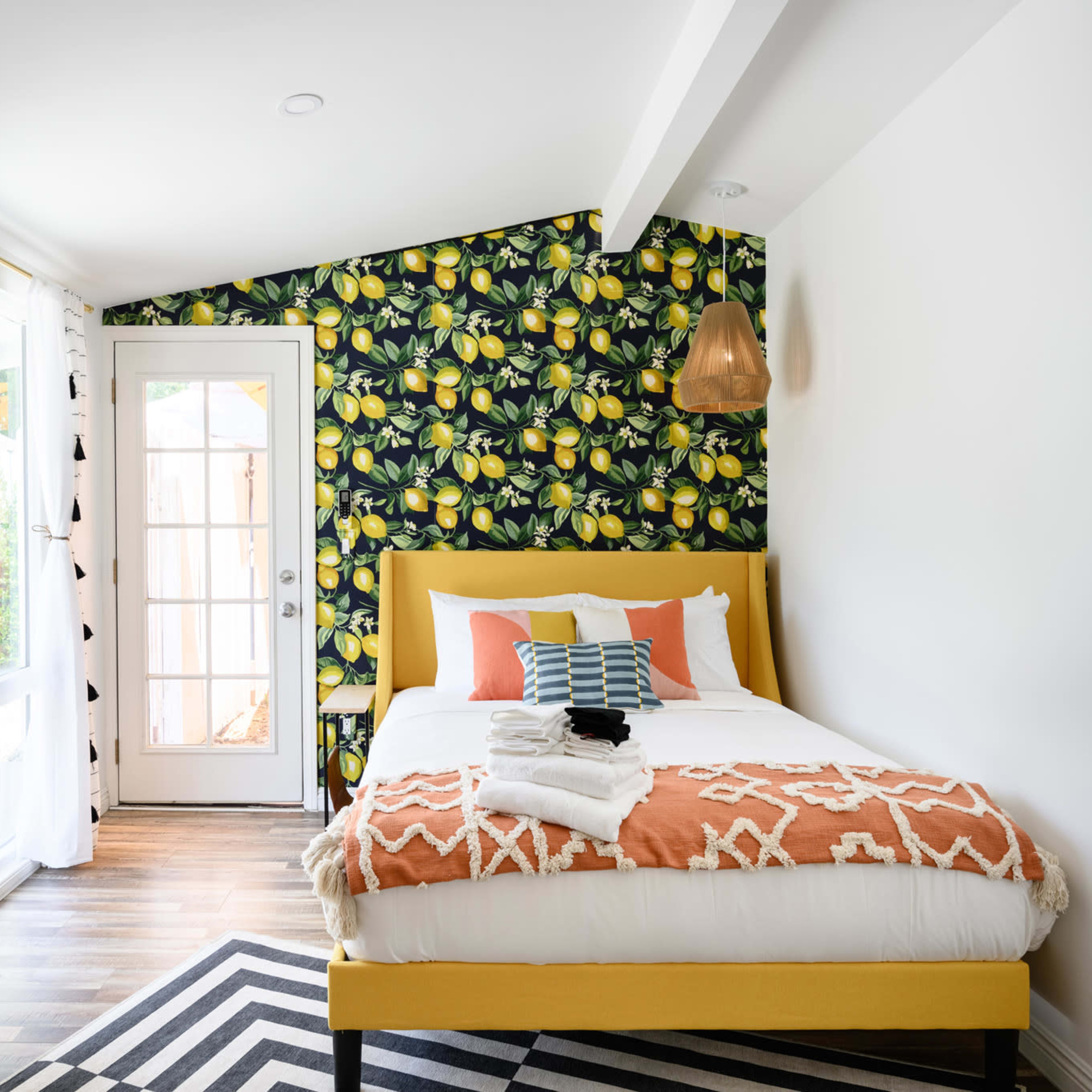 The room features a yellow bed with decorative pillows, a patterned lemon wallpaper, large windows, and stylish accent details.