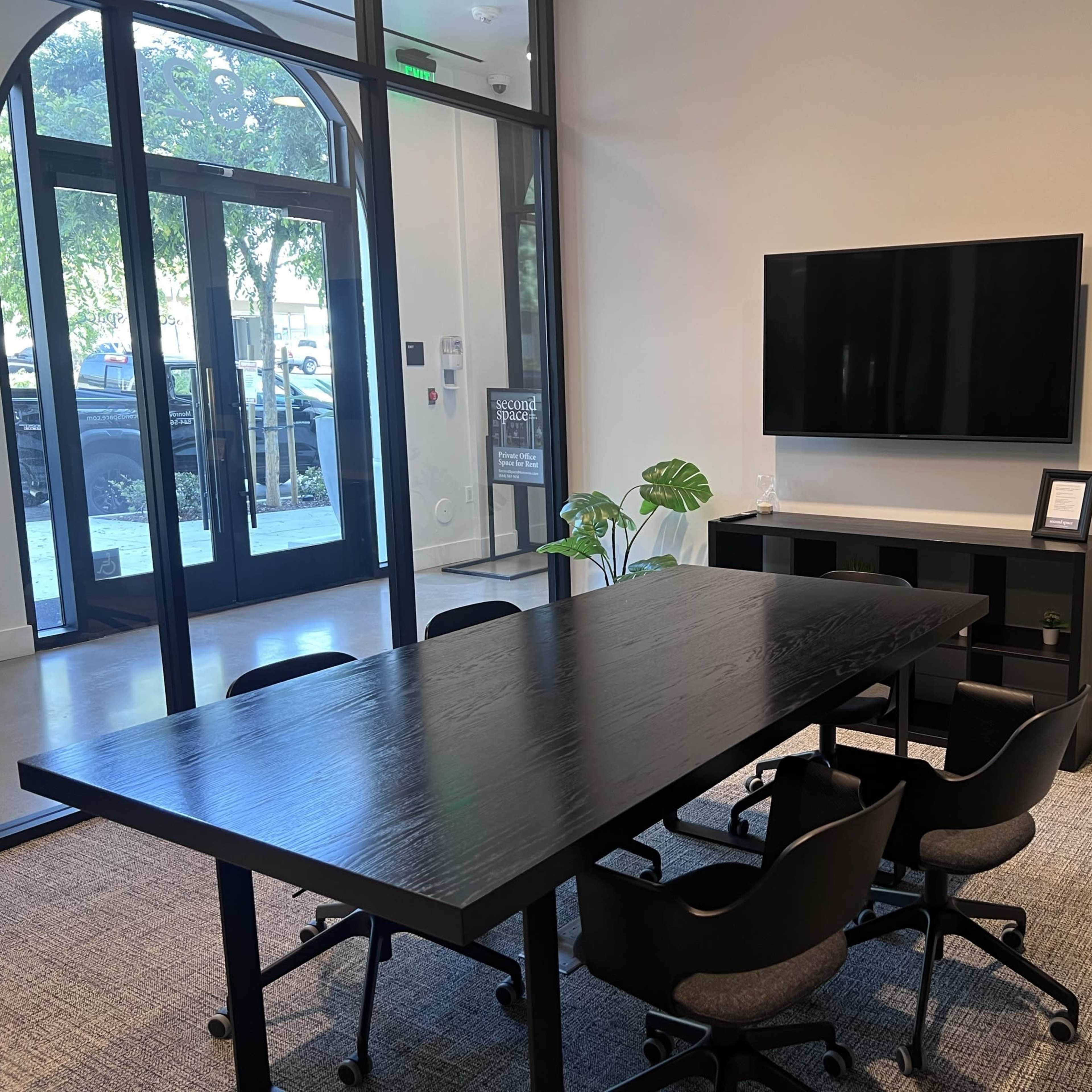 A modern conference room with a large black table, six black chairs, a wall-mounted television, and two potted plants.