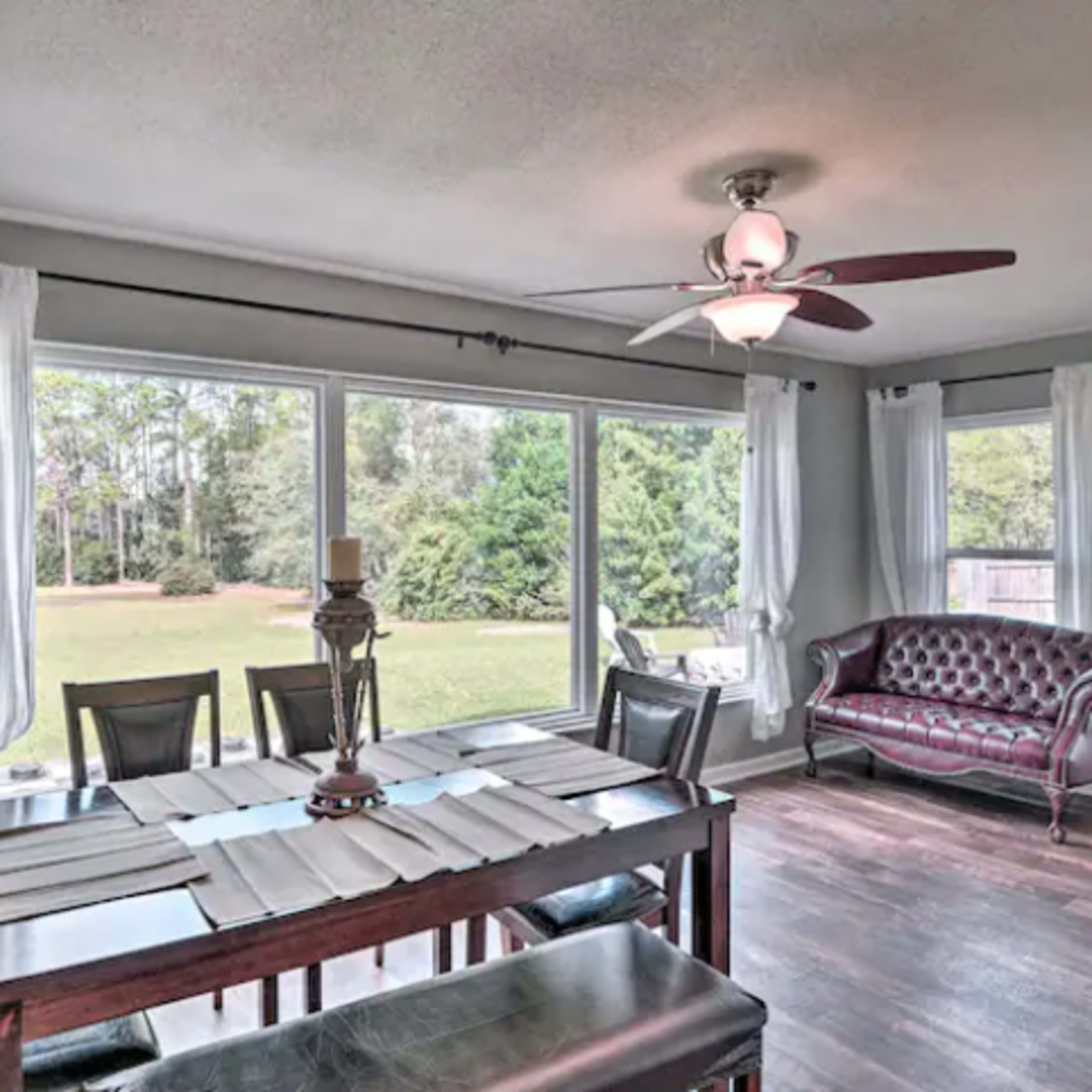 A dining area with a wooden table, eight chairs, and large windows overlooking a green outdoor space.